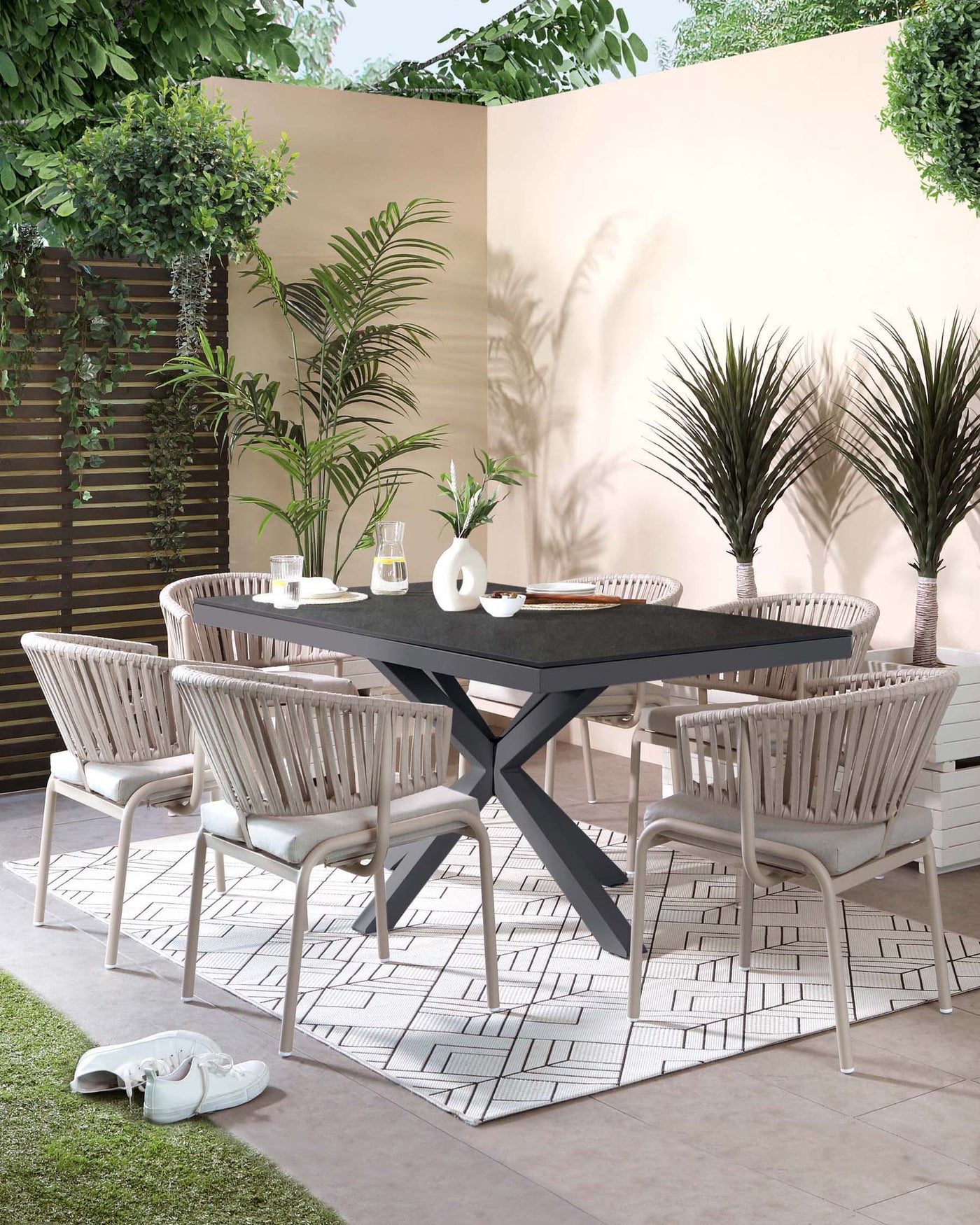 Outdoor dining set including a large, dark grey rectangle table with a unique X-shaped leg design, surrounded by six modern white armchairs with a vertical slat back design and light grey cushioned seats on an ornate white and grey geometric patterned area rug.