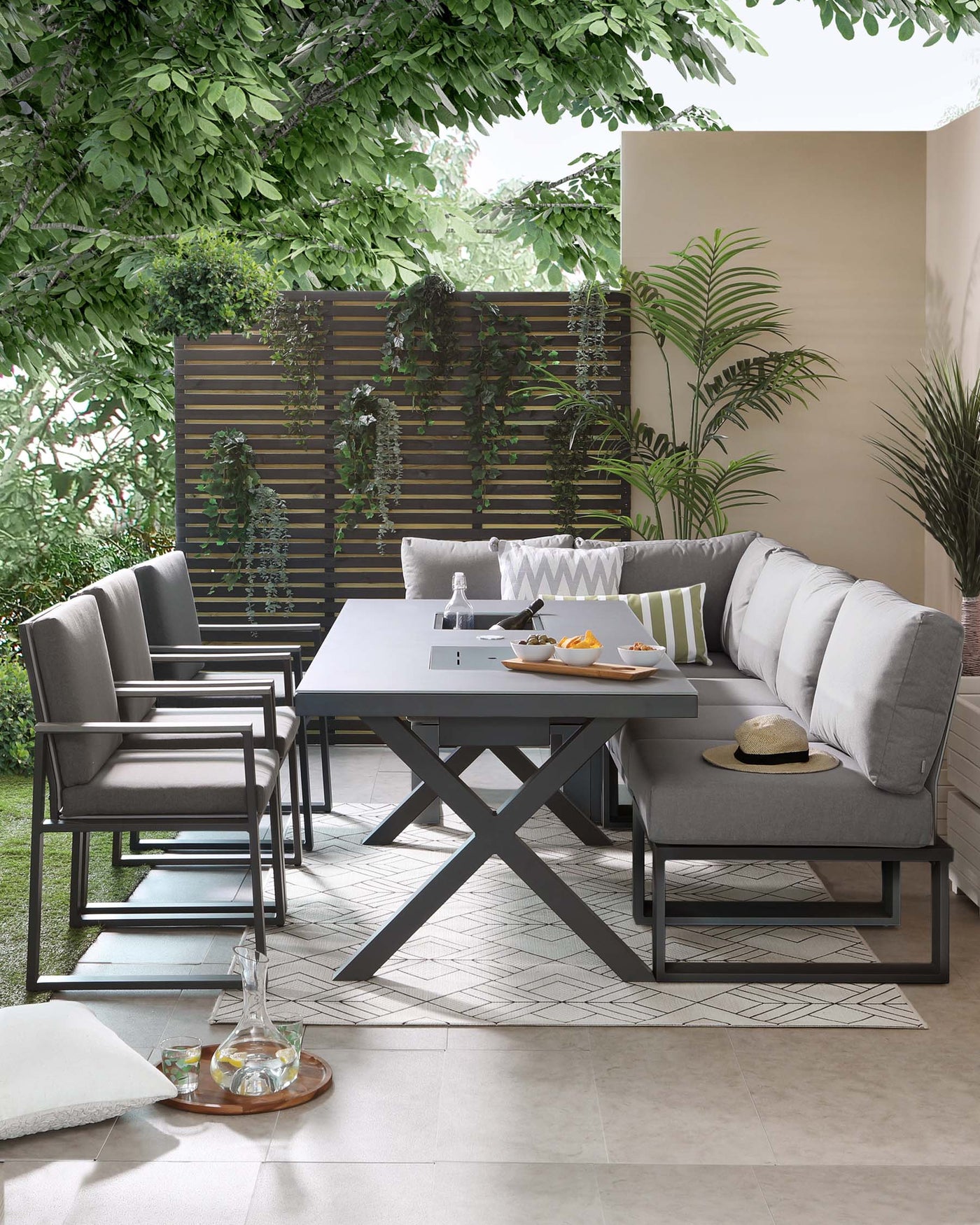 The image showcases a modern outdoor furniture set which includes a rectangular dining table with a sleek grey finish and a distinctive X-shaped base. Accompanied are four dining chairs and a lounge sofa with clean lines and a matching grey colour scheme. The chairs feature comfortable cushions and armrests, while the sofa is outfitted with plush backrest cushions for added comfort. The setting is complemented by green plants and trees, implying a tranquil garden environment.