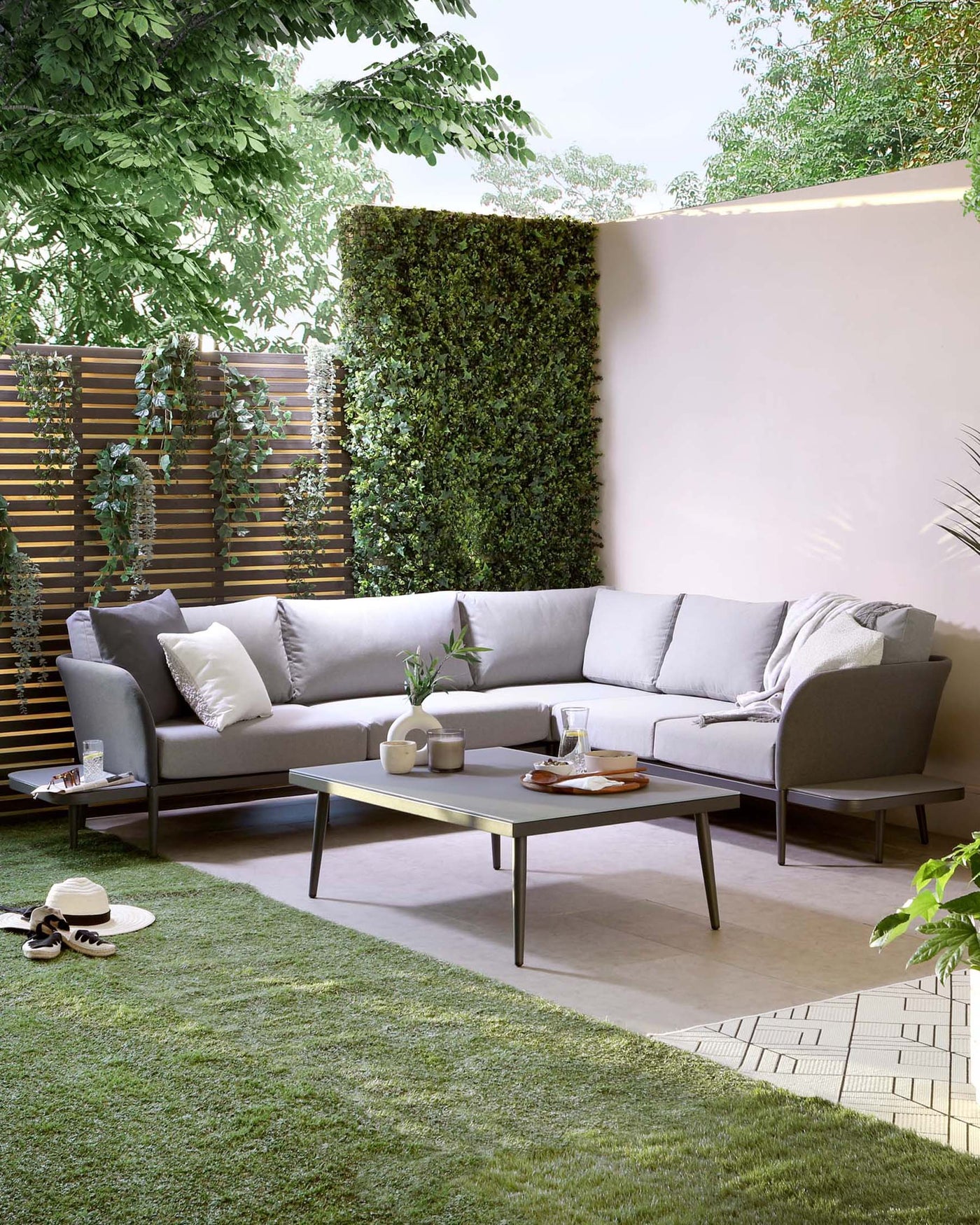 Modern outdoor furniture set featuring a sectional corner sofa with light grey cushions and a matching low-profile coffee table. The sofa is adorned with a variety of accent pillows in different shades of grey. The coffee table is rectangular with a smooth, matte finish and a slate grey colour, complementing the minimalist style of the seating. The furniture is arranged on a patio area with pavers and lush greenery in the background.