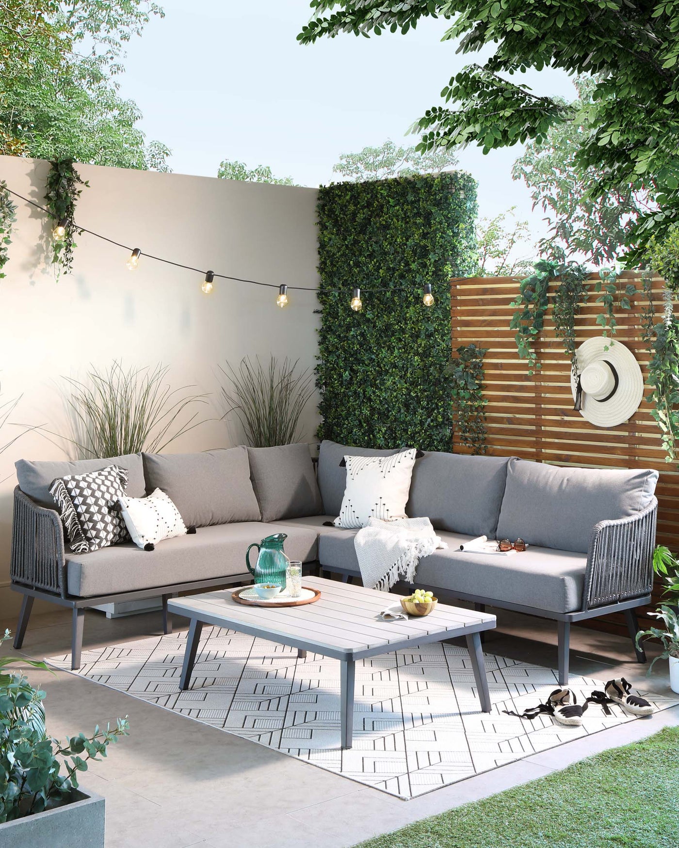 Modern outdoor furniture set featuring a sectional corner sofa with a sleek grey metal frame and plush light grey cushions, complemented by patterned throw pillows. A coordinating metal coffee table with a slatted top design sits at the centre. The set is arranged on a patterned outdoor rug, creating a cosy patio arrangement.