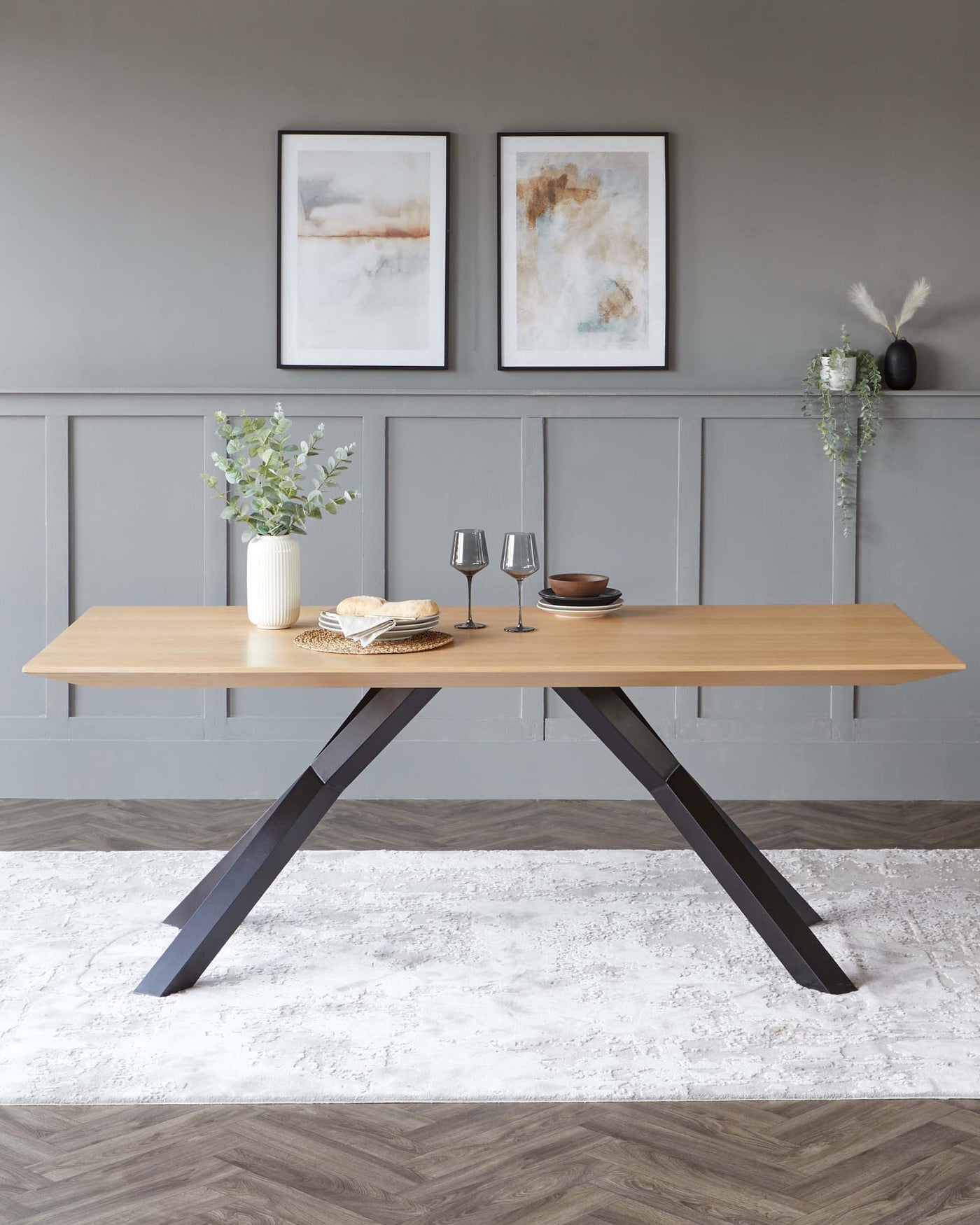 A modern rectangular dining table featuring a light wood tabletop and a pair of sturdy, angular black metal legs in an inverted V-shape, set upon a textured white area rug over herringbone wood flooring.