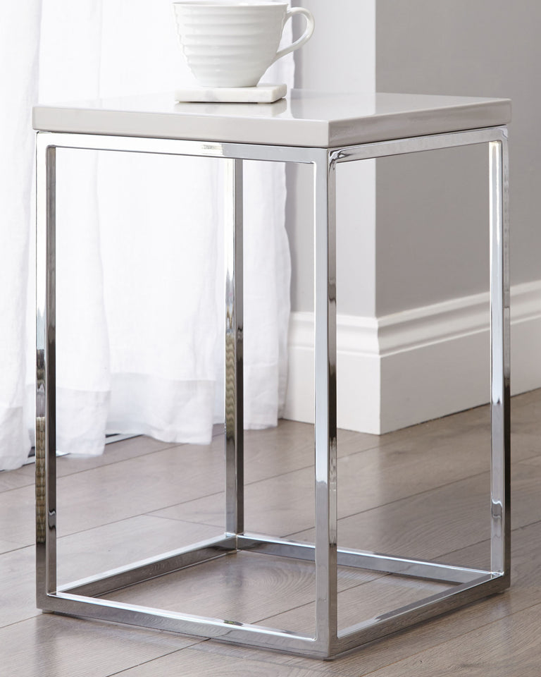 Modern minimalist square side table with a glossy white tabletop and a sleek, chrome-finished metal frame, accompanied by a white ceramic cup on a small saucer placed on top of the table.