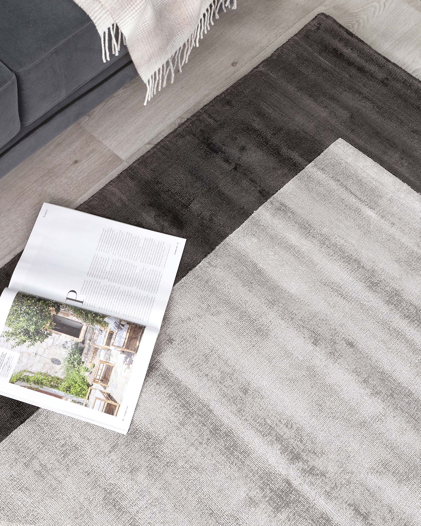 Modern ombre area rug transitioning from dark grey to light grey, complemented by a beige fringed throw blanket partially draped over a grey corner sofa, placed on a light wooden floor with an open magazine for a cosy, minimalist aesthetic.