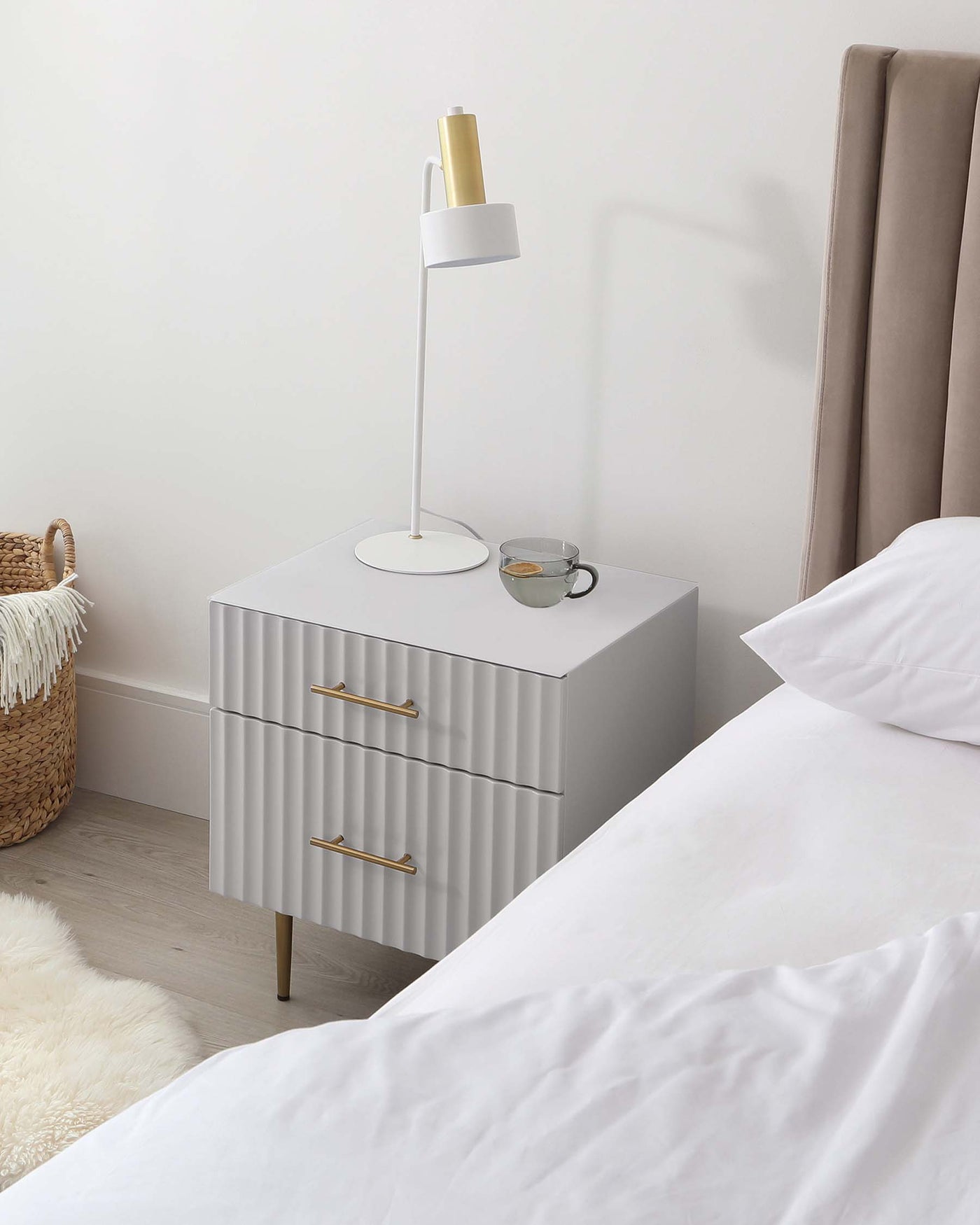 Modern light grey nightstand with two drawers featuring a sculpted, textured front and brass handles, complemented by four slender tapered legs with brass tips.