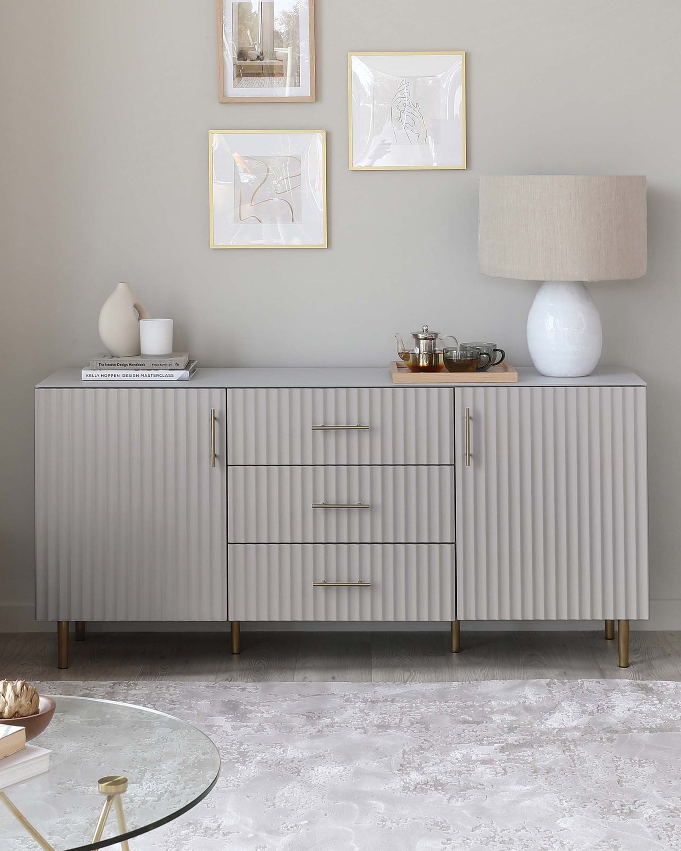Modern light grey sideboard with ribbed texture on doors and drawers, accented with sleek metallic handles and brass-finished tapered legs, accompanied by a simple round glass-top coffee table with a thin golden base positioned on an ornate light-coloured area rug.