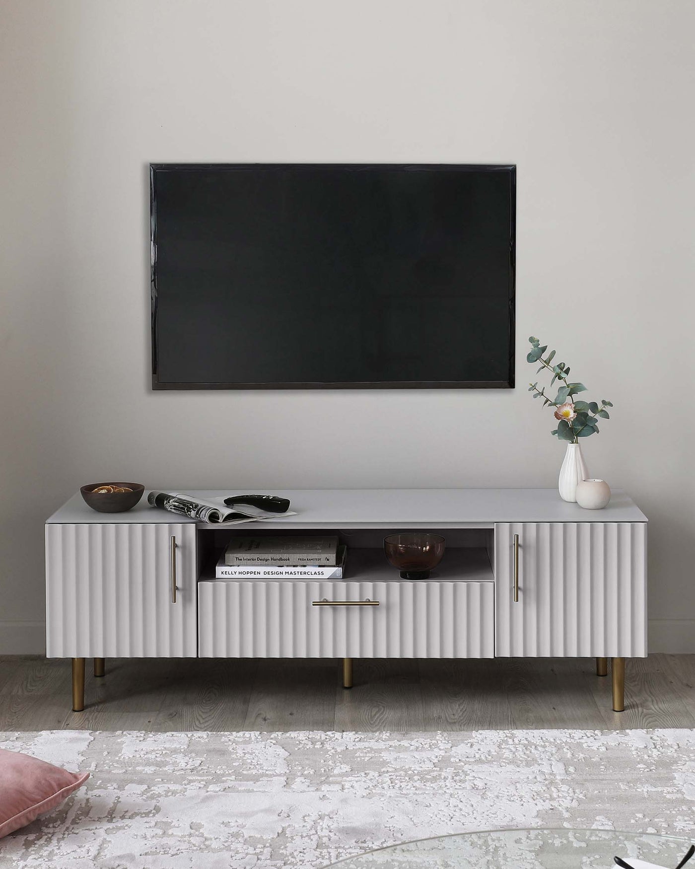 A contemporary white sideboard with a textured front, gold handles, and tapered wooden legs. The sideboard has two cabinet doors and a central shelf housing decorative items, positioned below a wall-mounted television, flanked by ornamental plant and books. A patterned light grey and white area rug is partially visible beneath it.
