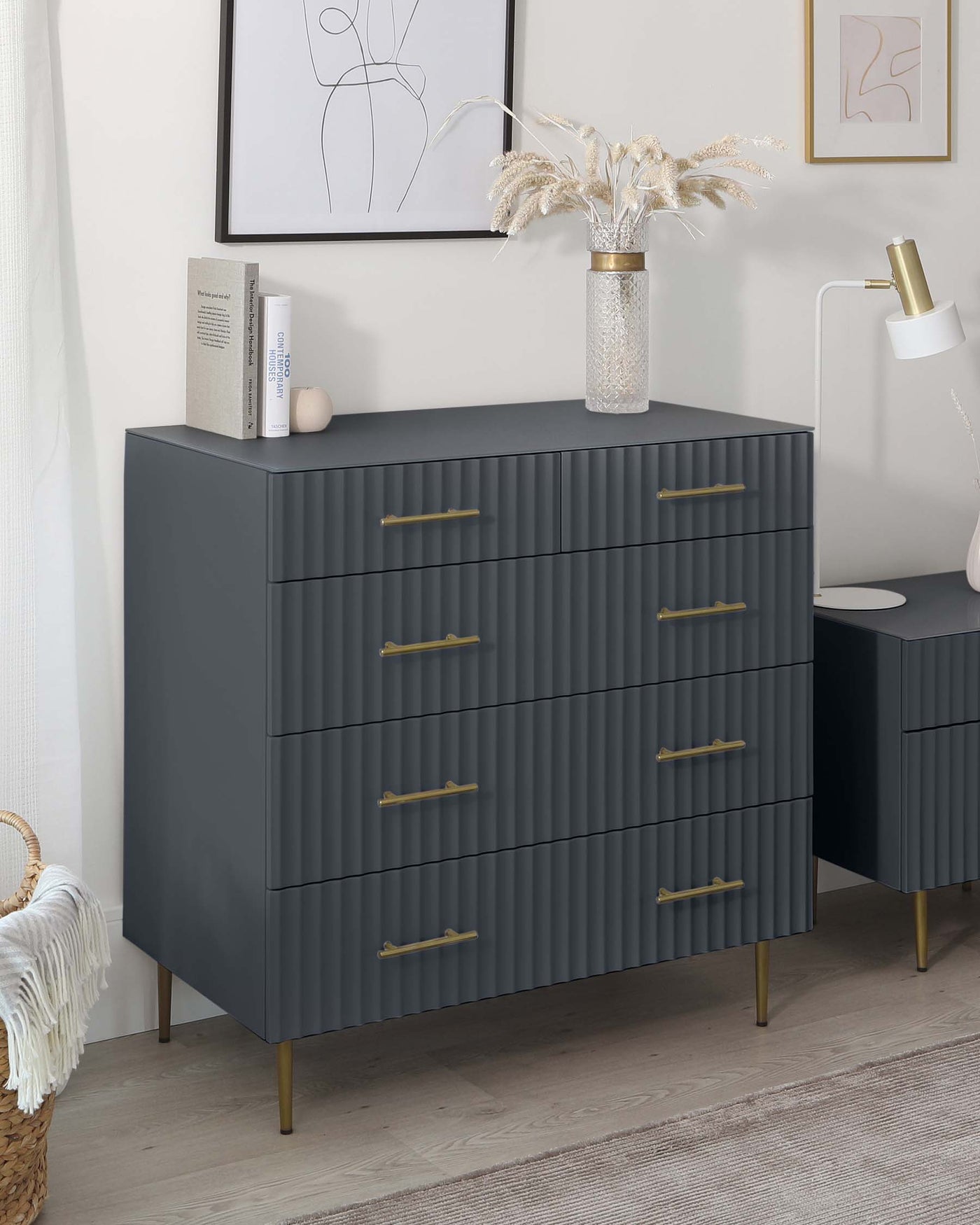 Modern matte grey six-drawer dresser with textured front panels and brass handles on tapered legs, accompanied by a matching desk with a white lamp, positioned against a white wall decorated with framed abstract art, next to a pampas grass vase and books.