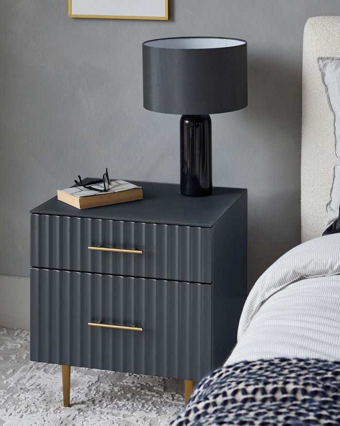 A contemporary charcoal grey nightstand with golden handles featuring three grooved drawers, a sleek dark tabletop, and tapered legs in a bedroom setting. A matching dark grey lamp with a large round shade, a book, and a pair of glasses are placed on top of the nightstand, complementing the modern aesthetic.