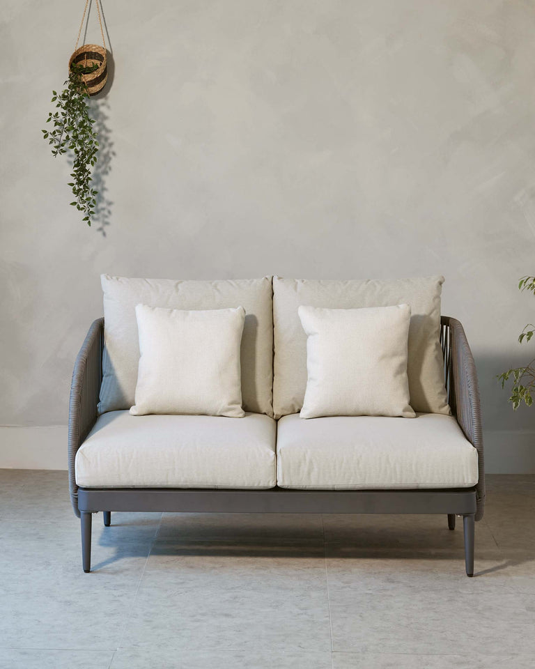 Modern two-seater sofa with a sleek, dark wooden frame and light beige cushions, accompanied by four matching throw pillows. The sofa is set against a neutral wall with a single hanging potted plant to the left.