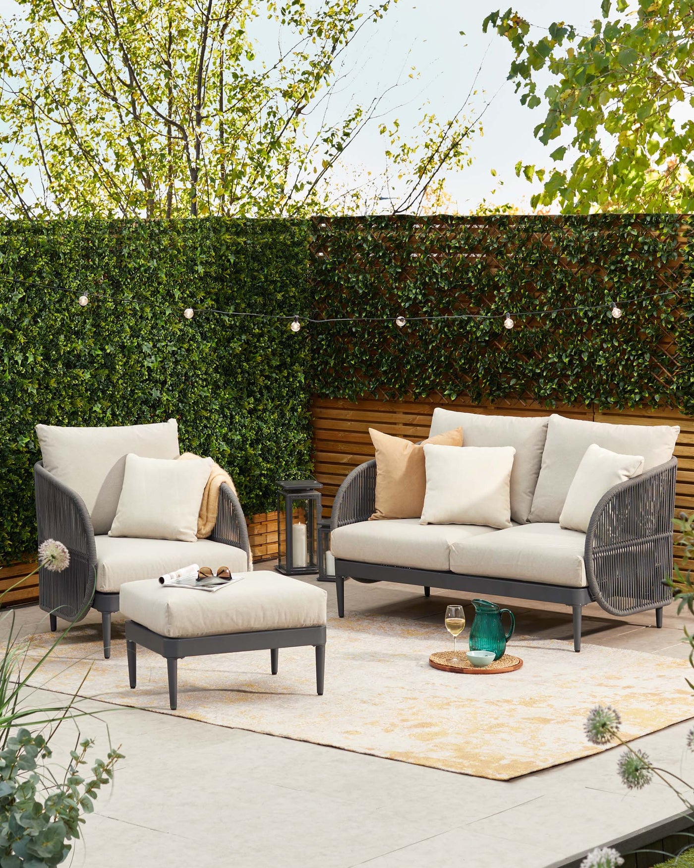 Elegant outdoor furniture set featuring a three-seater sofa with plush beige cushions, two high-back armchairs with matching cushions, and a rectangular ottoman, all with dark grey frames and woven side detailing. A small wooden side table and patterned area rug complement the arrangement.