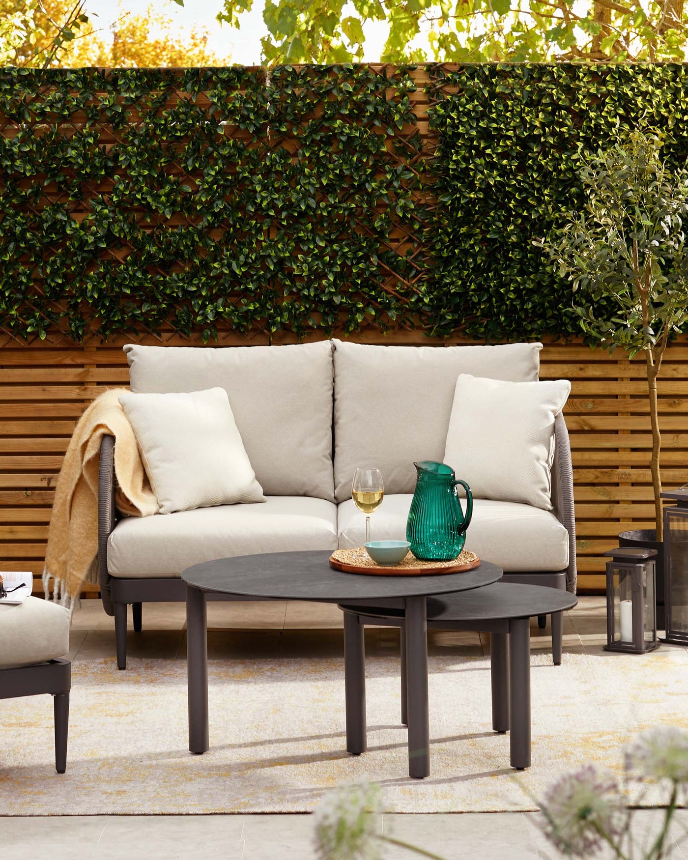 Outdoor patio lounge set with a three-seat sofa featuring neutral-toned cushions and a plush throw, paired with two round nesting coffee tables in a matte black finish.