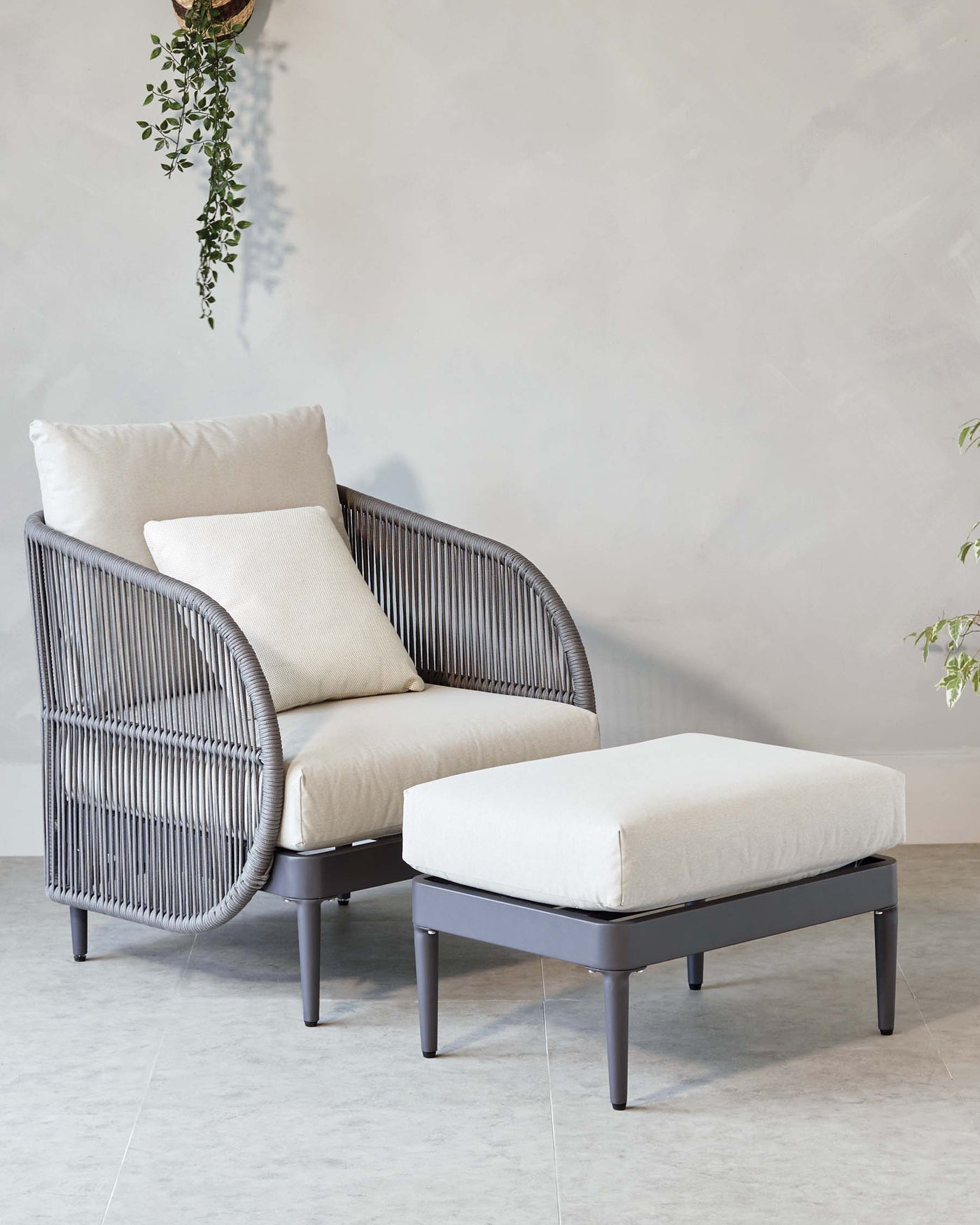 Modern armchair with taupe vertical wicker design and cream cushions paired with a matching simple square ottoman on a clean, neutral tiled floor, accented with a hanging potted plant with trailing green leaves.