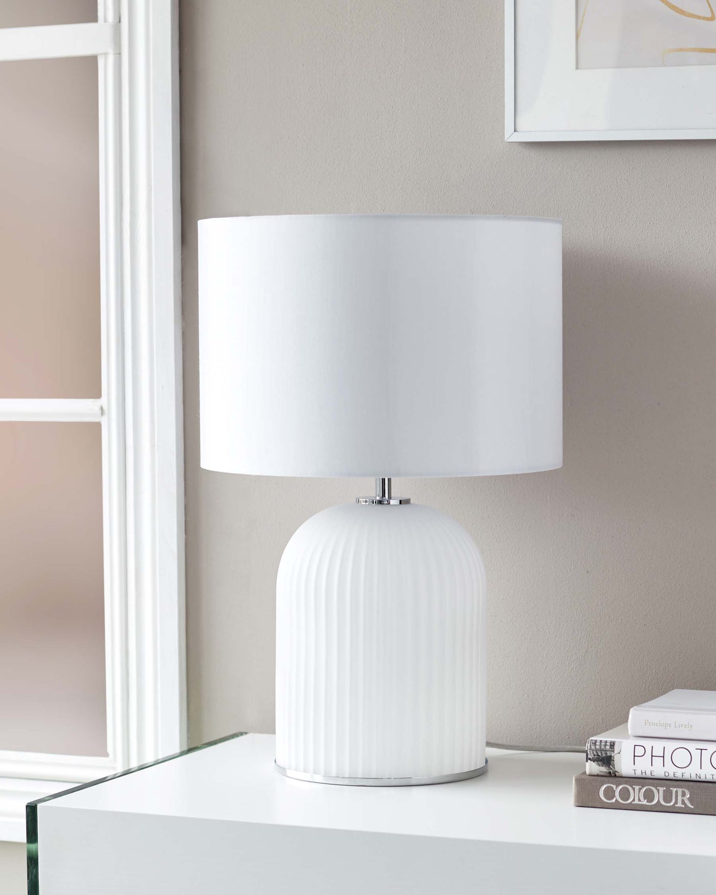 Elegant white table lamp with a cylindrical, ribbed base and a smooth, wide drum shade, displayed on a sleek, reflective glass table.