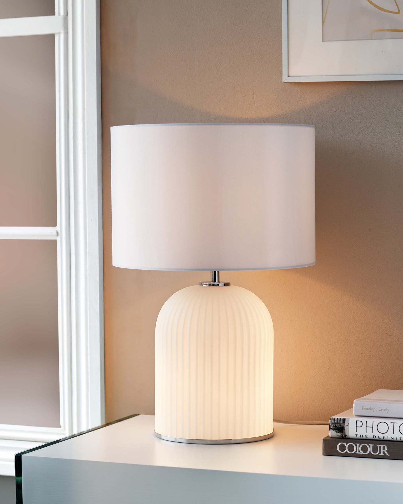 Modern table lamp with a ribbed ceramic base and a large cylindrical white shade, placed on a glass table next to books, with artwork on the wall and a window in the background.