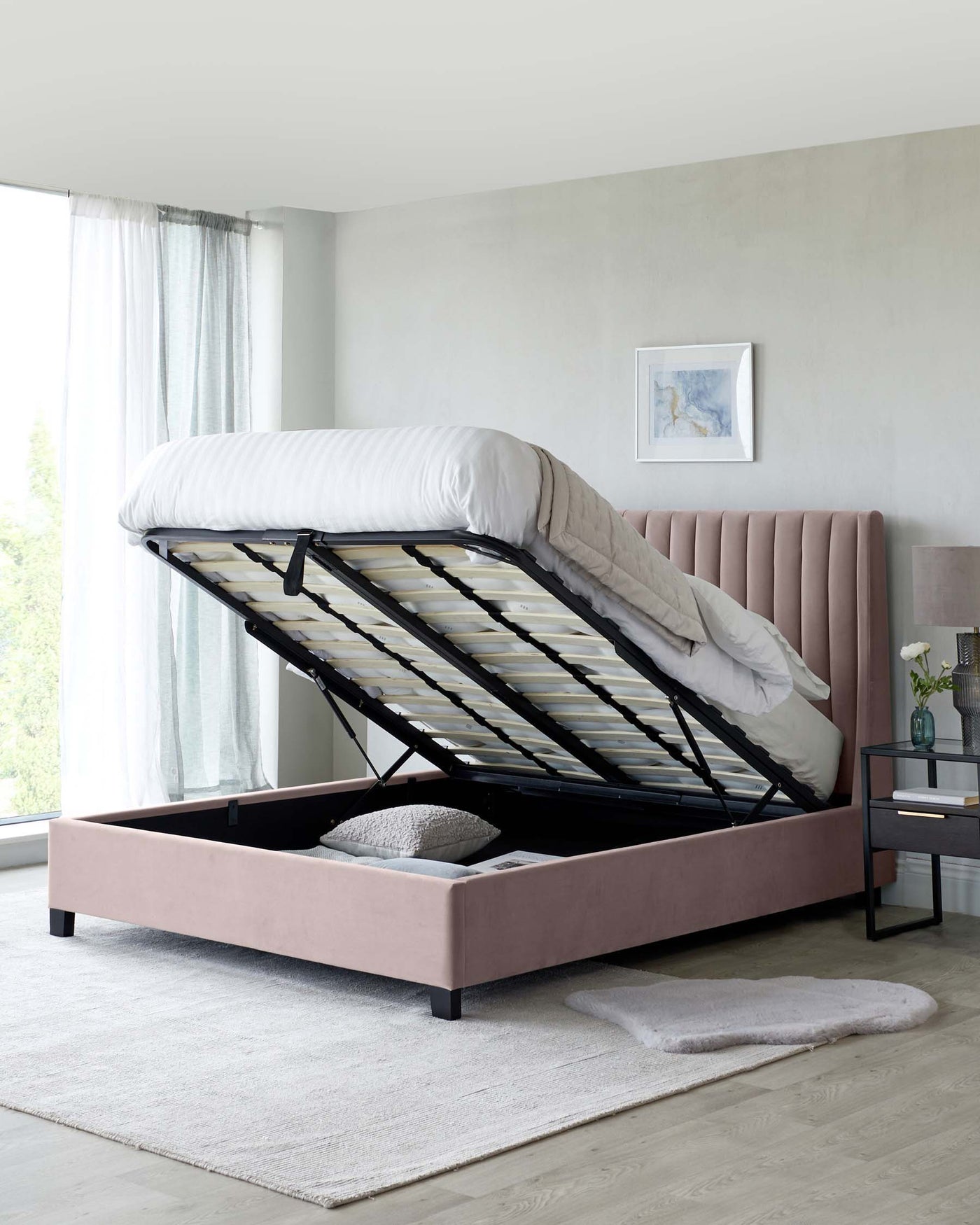 Upholstered storage bed with lifted mattress frame revealing under-bed storage space, featuring a plush, channel-tufted headboard in a soft blush colour, accented with dark wooden legs, set against a neutral bedroom decor.