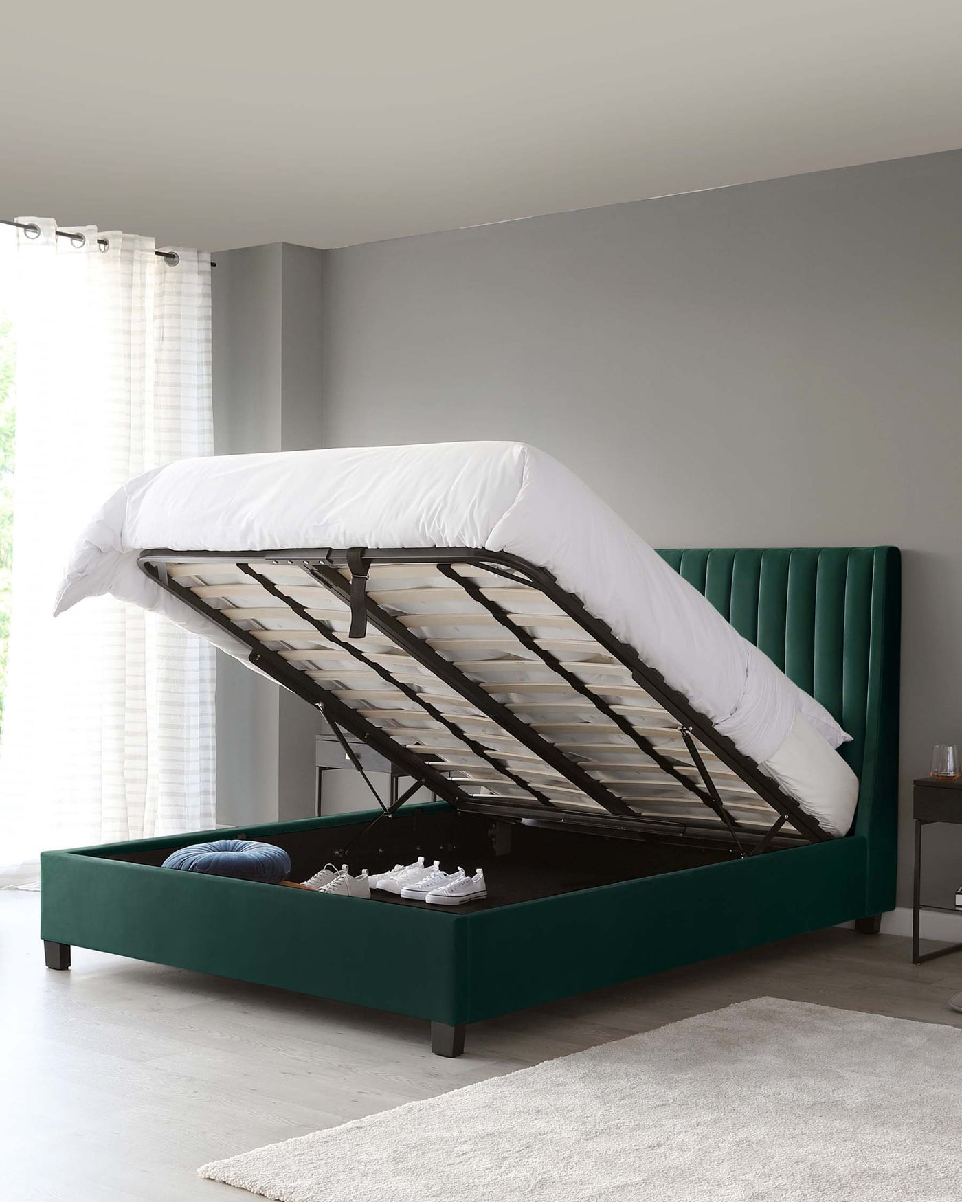 An elegant emerald green upholstered storage bed with a lifted frame revealing a spacious under-mattress compartment. The bed features a tufted, padded headboard and a low-profile foot and side rails on dark wooden legs. A white duvet partly covers the slatted base.