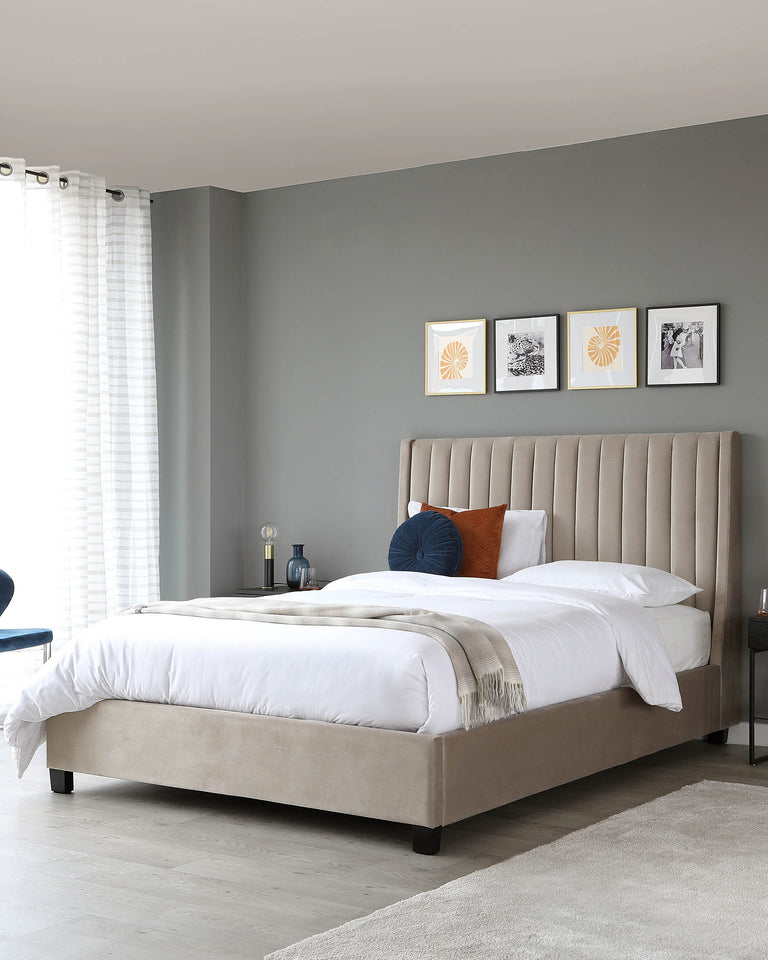 Elegant beige upholstered bed frame with a high tufted headboard, flanked by a small black side table with a contemporary lamp, and a round blue ottoman at the foot.