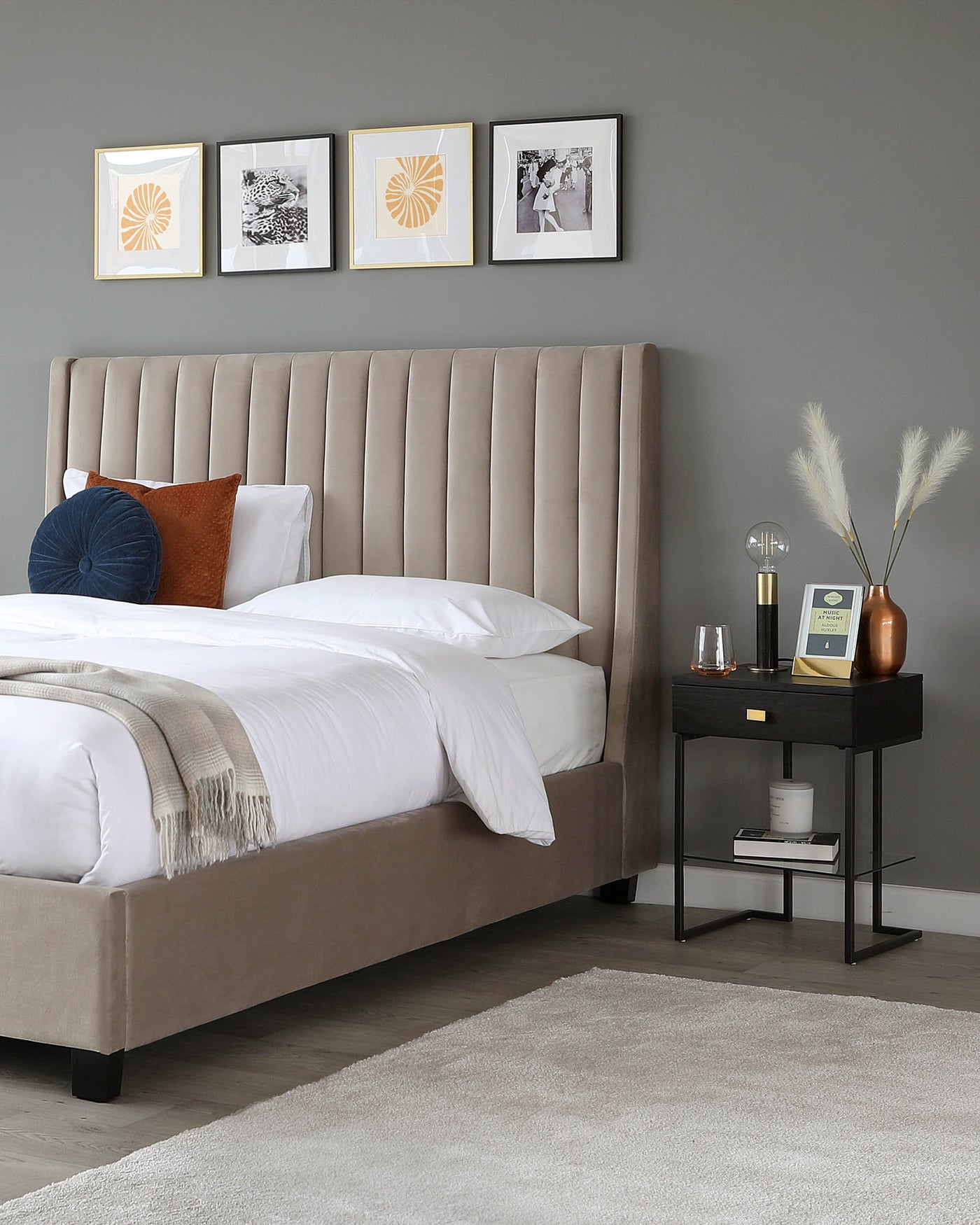 A plush, taupe upholstered bed with a high, vertically channel-tufted headboard and a thin neutral-toned blanket at the foot. Beside the bed is a modern black nightstand with clean lines, featuring an open shelf and a drawer. A small table lamp with a brass finish, decorative vases, and a book are placed on top of the nightstand. A soft grey area rug lies on the floor, partially under the bed.