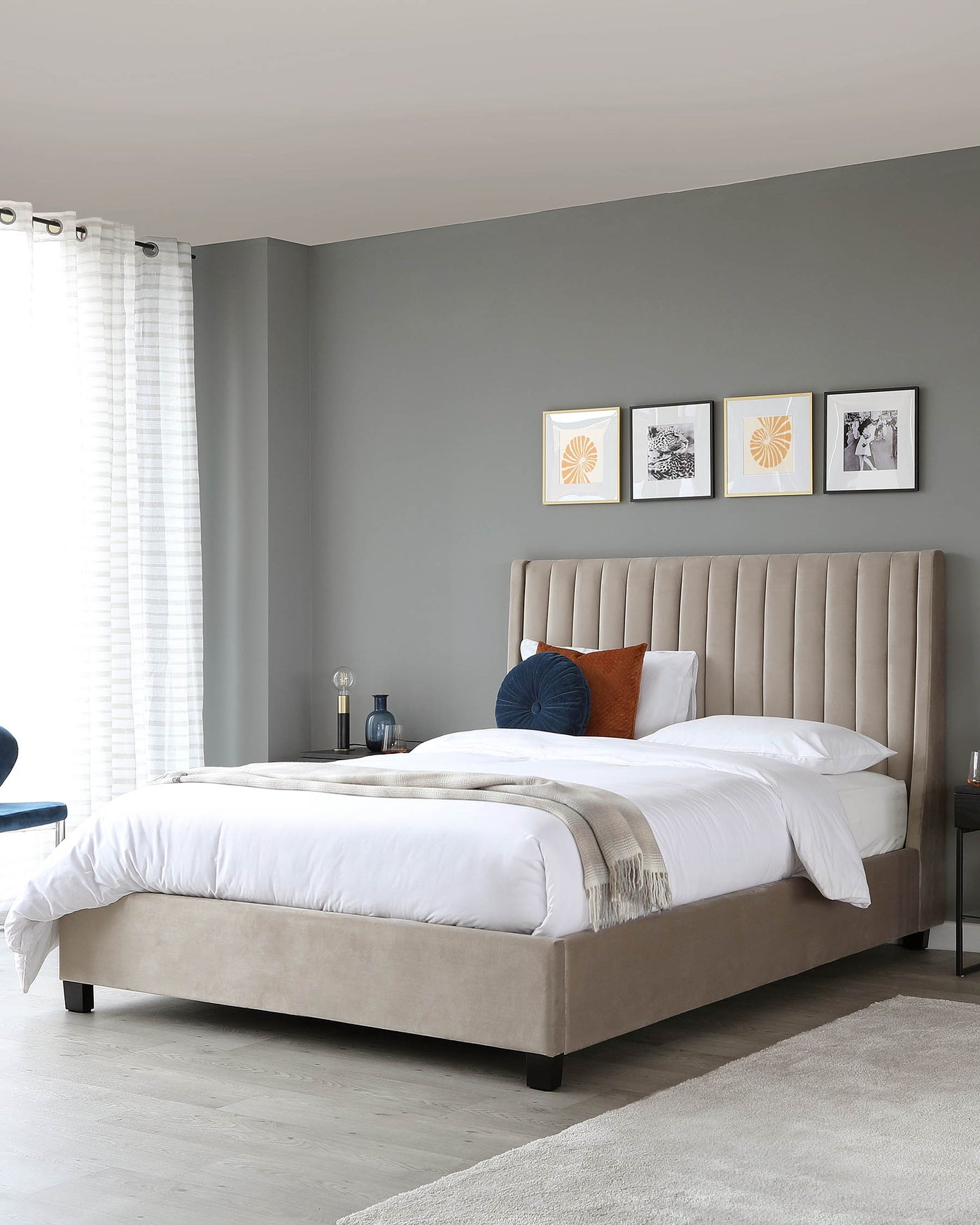 Elegant modern bedroom featuring a velvet upholstered king-sized platform bed with a high headboard displaying vertical channel tufting. The bed is layered with crisp white bedding, a beige throw, and assorted decorative pillows. To the side, a small nightstand with a contemporary table lamp is visible. No other furniture is prominently displayed.