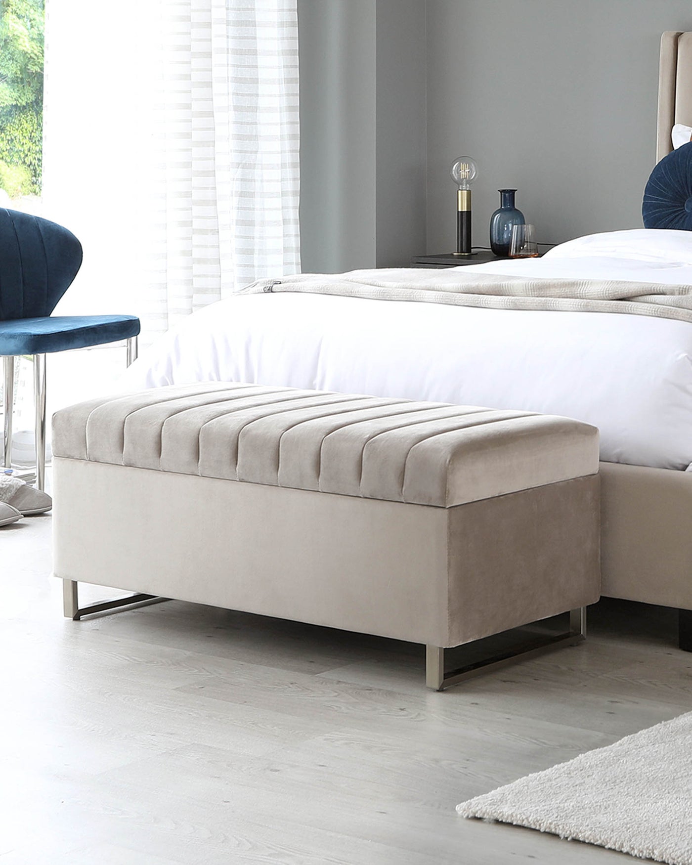 Elegant light grey upholstered bench with a plush, tufted top cushion, featuring a streamlined design and metallic legs, positioned at the foot of a bed in a modern bedroom setting.