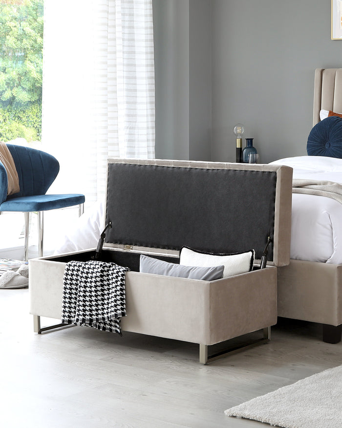 Beige upholstered storage bench with a tufted grey interior, situated at the foot of a bed, with metallic legs and a houndstooth throw blanket draped over one corner.