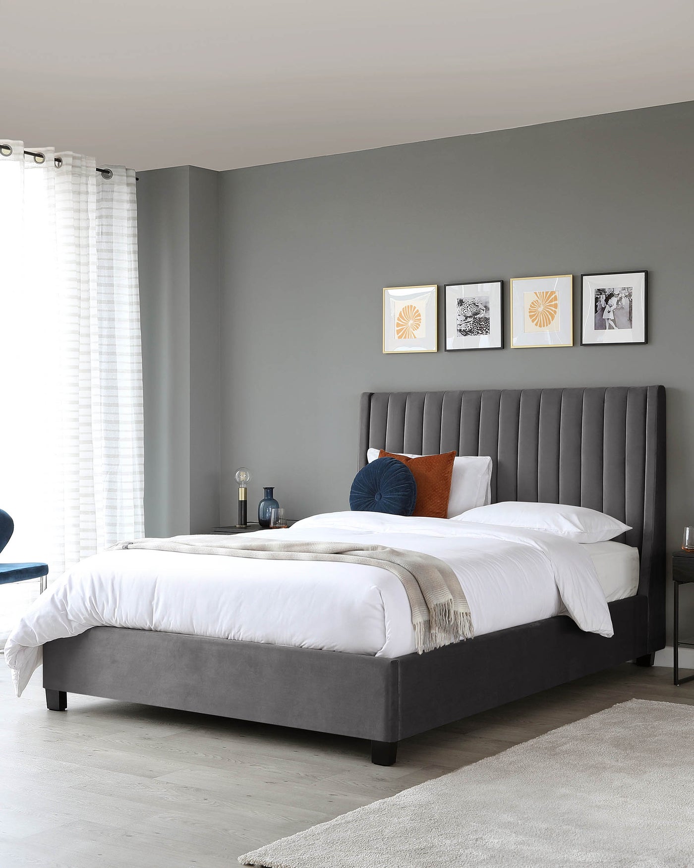 A modern grey upholstered platform bed with a high vertical channel-tufted headboard, complemented by white bedding, two pillows, and a beige fringed throw blanket. A small black nightstand with a table lamp is beside the bed, and a round blue chair is seen partially in the corner. The room features a light grey floor and area rug with a smooth texture.