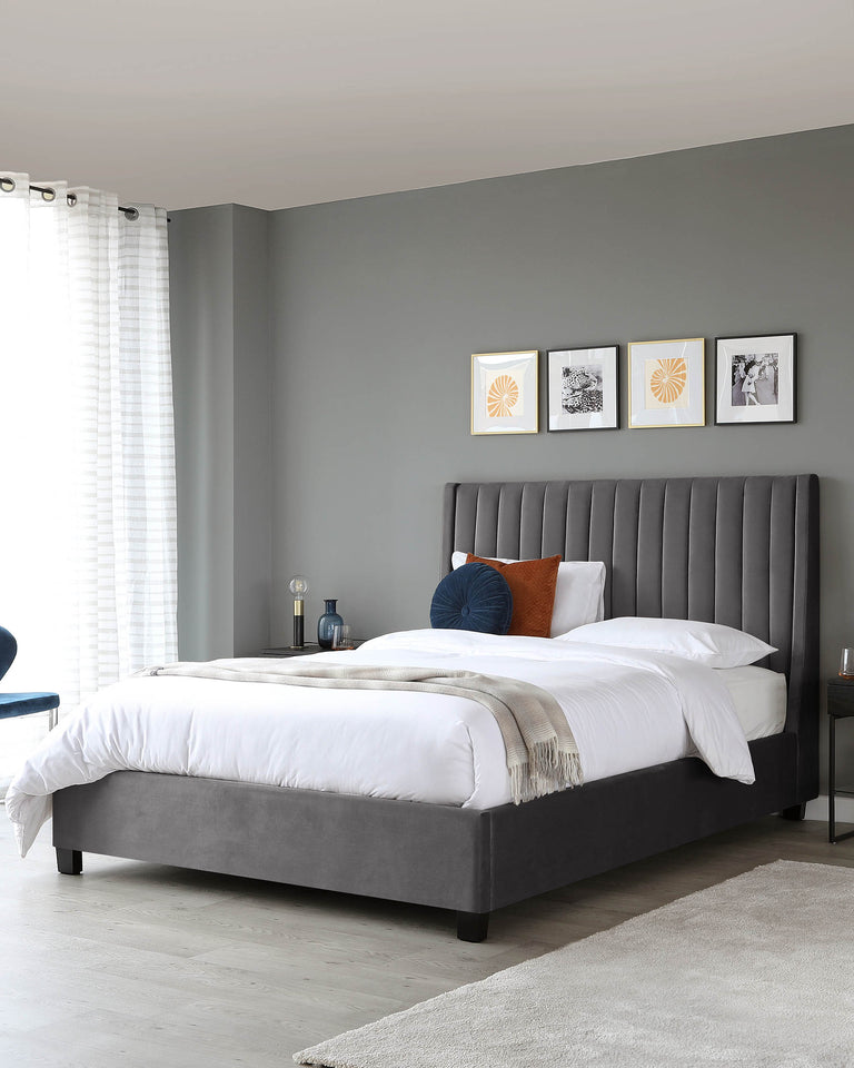 Elegant grey upholstered bed with a tall headboard featuring vertical channel tufting, complemented by white bedding, a beige throw, and decorative pillows in navy and rust tones. Two dark nightstands flank the bed, with minimalistic design lamps atop each. The room has a modern and serene aesthetic.