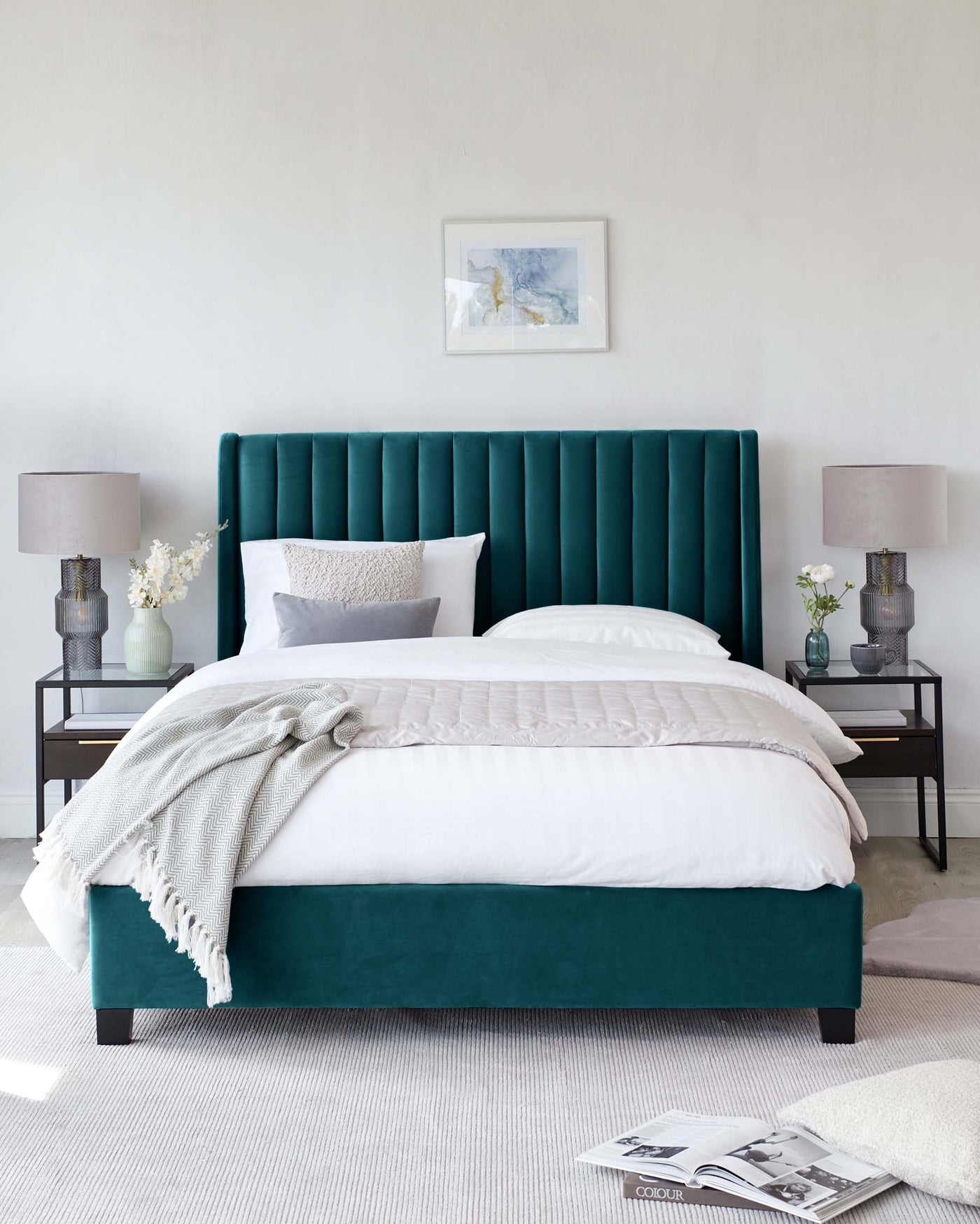 An elegant emerald green upholstered bed with a tall, vertical channel-tufted headboard, flanked by two black metal side tables with glass tops, each adorned with a grey lamp and white floral arrangement.