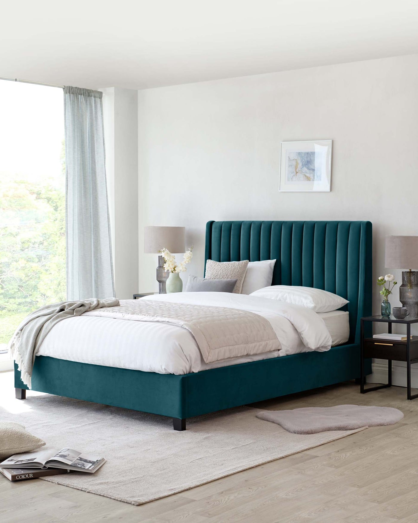 Elegant bedroom setup featuring a teal upholstered bed with a vertically channelled headboard and a matching bed frame. The bed is dressed with crisp white linens and assorted neutral pillows. Beside the bed is a sleek, black nightstand with an open shelf and a drawer, topped with a modern silver lamp and decorative vases. The room is anchored by a soft beige area rug, and a grey throw is casually draped over the foot of the bed.
