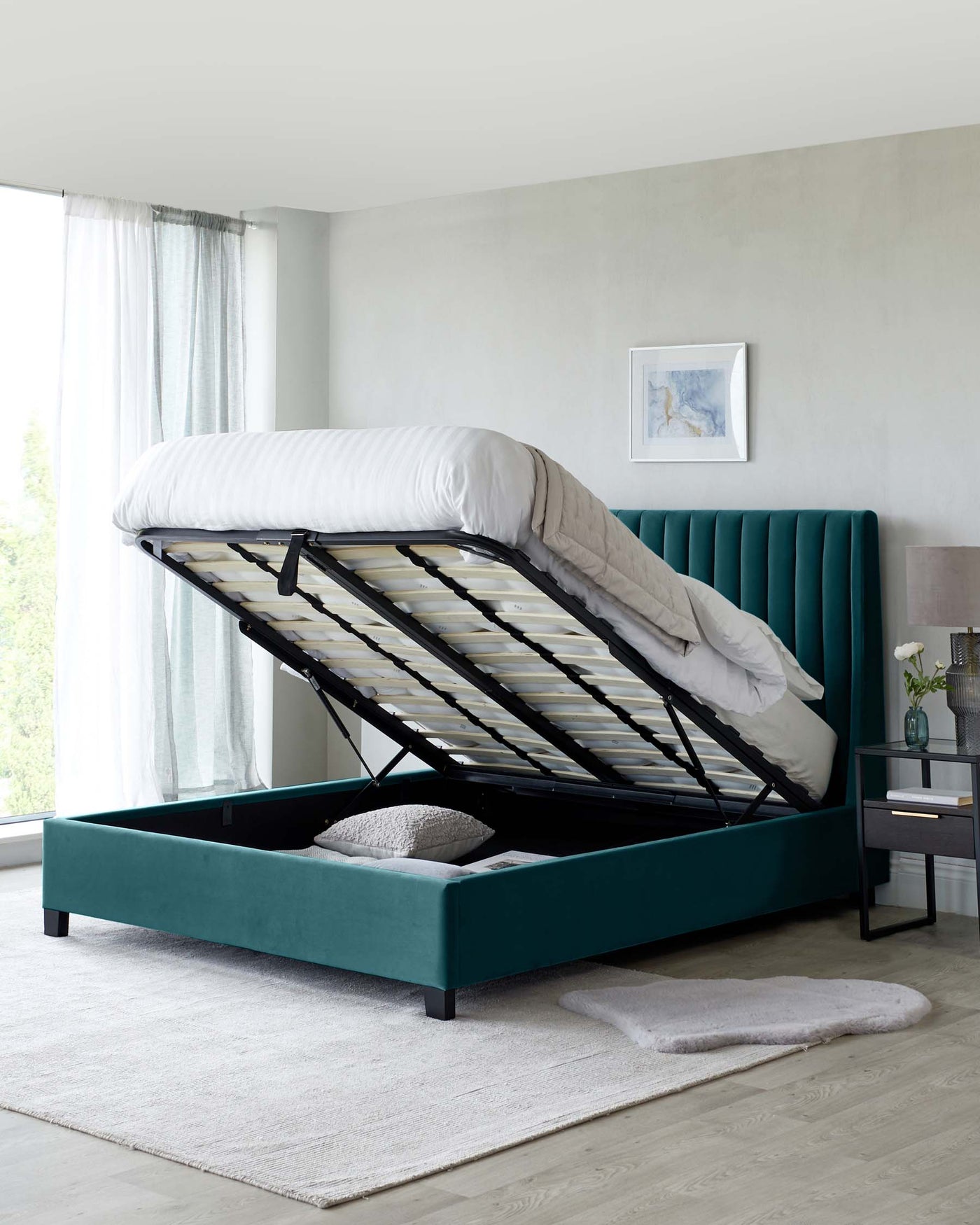 Elegant teal upholstered storage bed with a vertically channelled headboard and an open frame revealing a spacious under-mattress storage area. The bed is complemented by a black and wood accent nightstand with a lamp and floral arrangement, juxtaposed against a subdued room decor with grey walls and light wood flooring.
