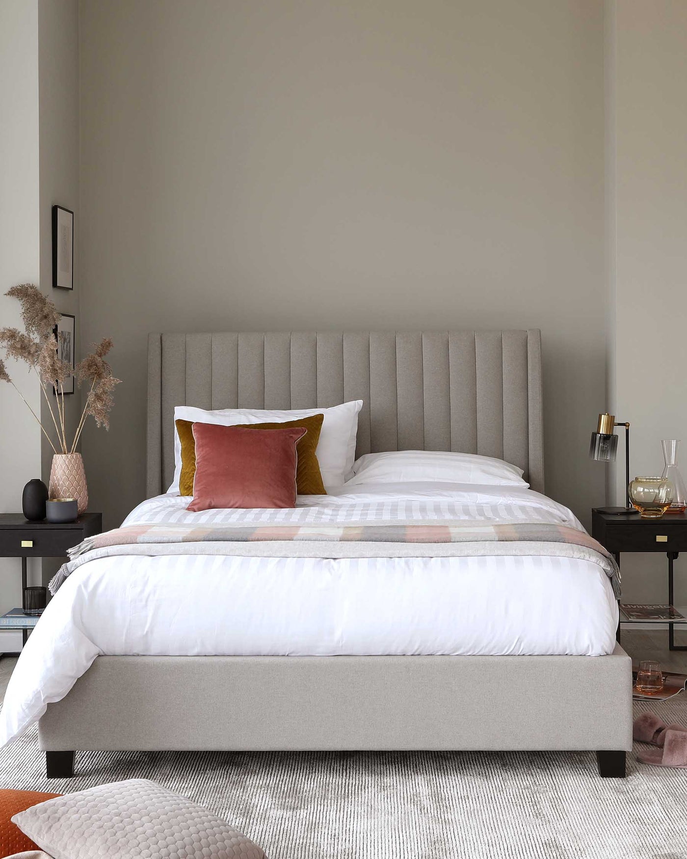 Contemporary bedroom showcasing a sleek upholstered platform bed with a tall, channel-tufted headboard in a neutral grey fabric. The bed is dressed in white bedding with a checkered pattern and accented with a mustard and a rust-coloured pillow. A simple black nightstand with an open shelf is placed beside the bed, holding a small table lamp, books, and decorative items.
