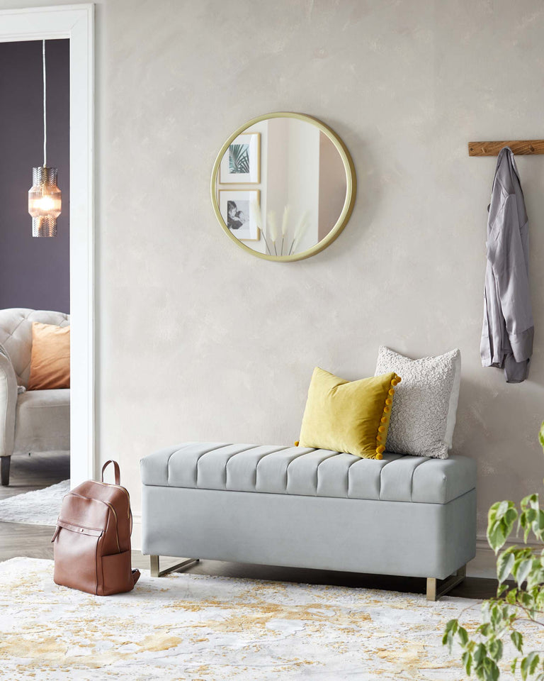 Elegant light grey upholstered bench with tufted cushion top, resting on minimalist metallic legs. The bench is accessorized with two decorative pillows, one in a mustard yellow colour with a pom-pom edge and the other in a textured grey fabric.