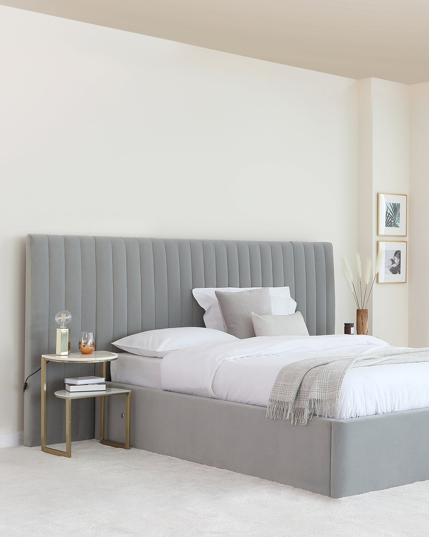 Modern minimalist bedroom furniture featuring a large king-size bed with an upholstered grey headboard in a vertical channel-tufted design. The bed is dressed in white linens, accompanied by a sleek, gold-finished metal side table with a circular top and a smaller rectangular lower shelf. The side table is accessorized with a couple of books, a decorative glass bottle, and a glass with a beverage. The room's decor is complemented by neutral tones and a couple of framed wall art pieces above the side table.