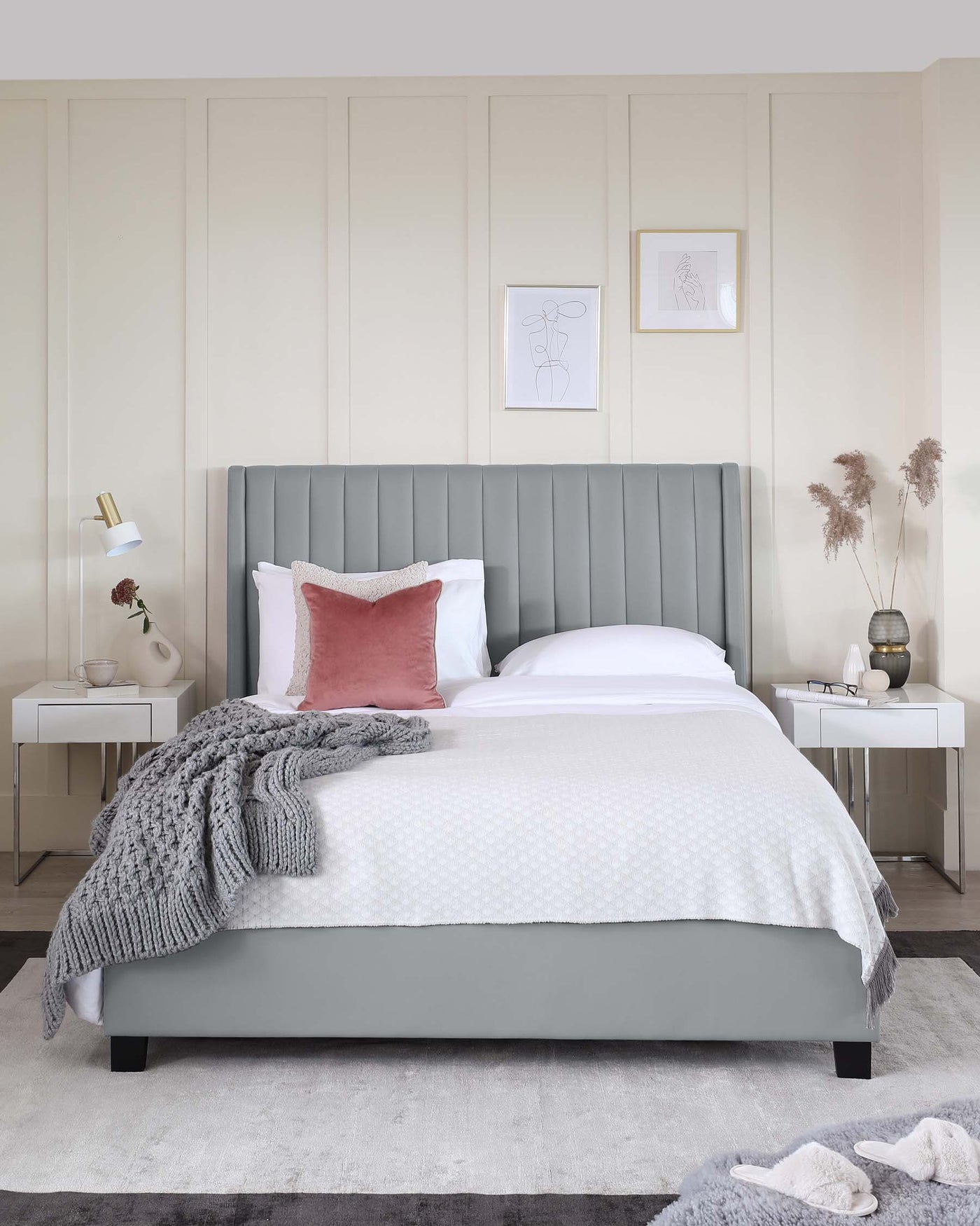 Modern bedroom furniture set featuring a large grey upholstered bed with a vertical channel tufted headboard and a white duvet. Flanking the bed are two matching white nightstands with table lamps, accessorized with decorative vases. The furniture stands on a textured grey area rug over a light wooden floor.