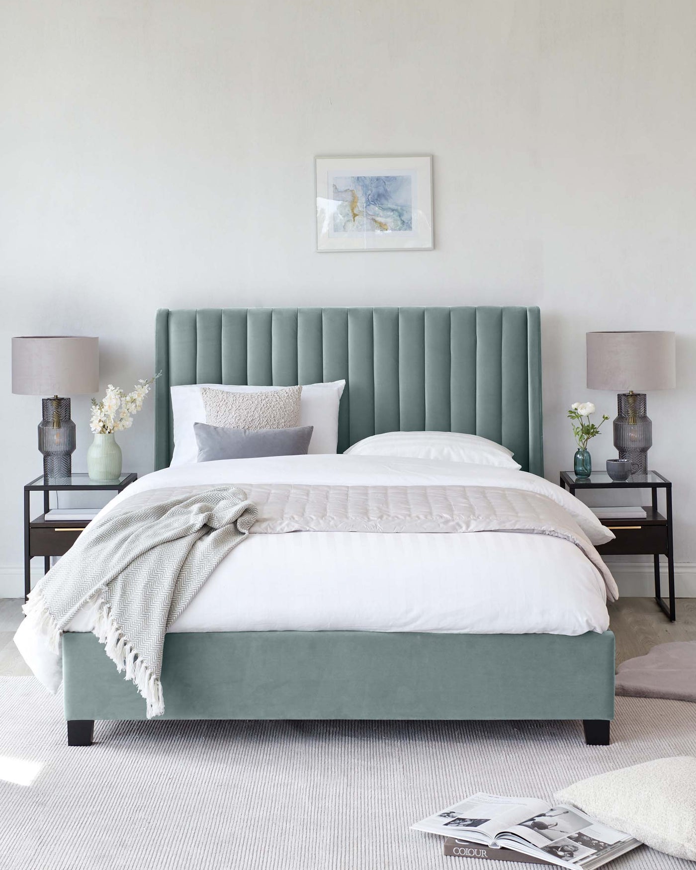 Elegant bedroom setup featuring an upholstered king-size bed with a tall, tufted headboard in a soothing green hue. Two matching nightstands with a modern, minimal design flank the bed, each bearing a stylish table lamp with a textured base and white shade. The bed is dressed in crisp white linens, accented by a textured throw and decorative pillows. A simple framed artwork hangs above the headboard, and the room is softened by a neutral area rug underfoot.