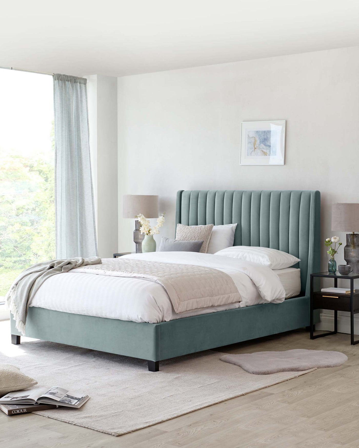Elegant bedroom featuring a large king-size bed with a teal upholstered headboard displaying vertical channel tufting. The bed is dressed in white and beige bedding with a plush white comforter and assorted throw pillows. Flanking the bed are two nightstands, one with a modern table lamp and a vase of white flowers, the other with a glass vase and books. The serene setting is complemented by a light grey area rug beneath the bed and soft curtains framing a window with a view of greenery outside.