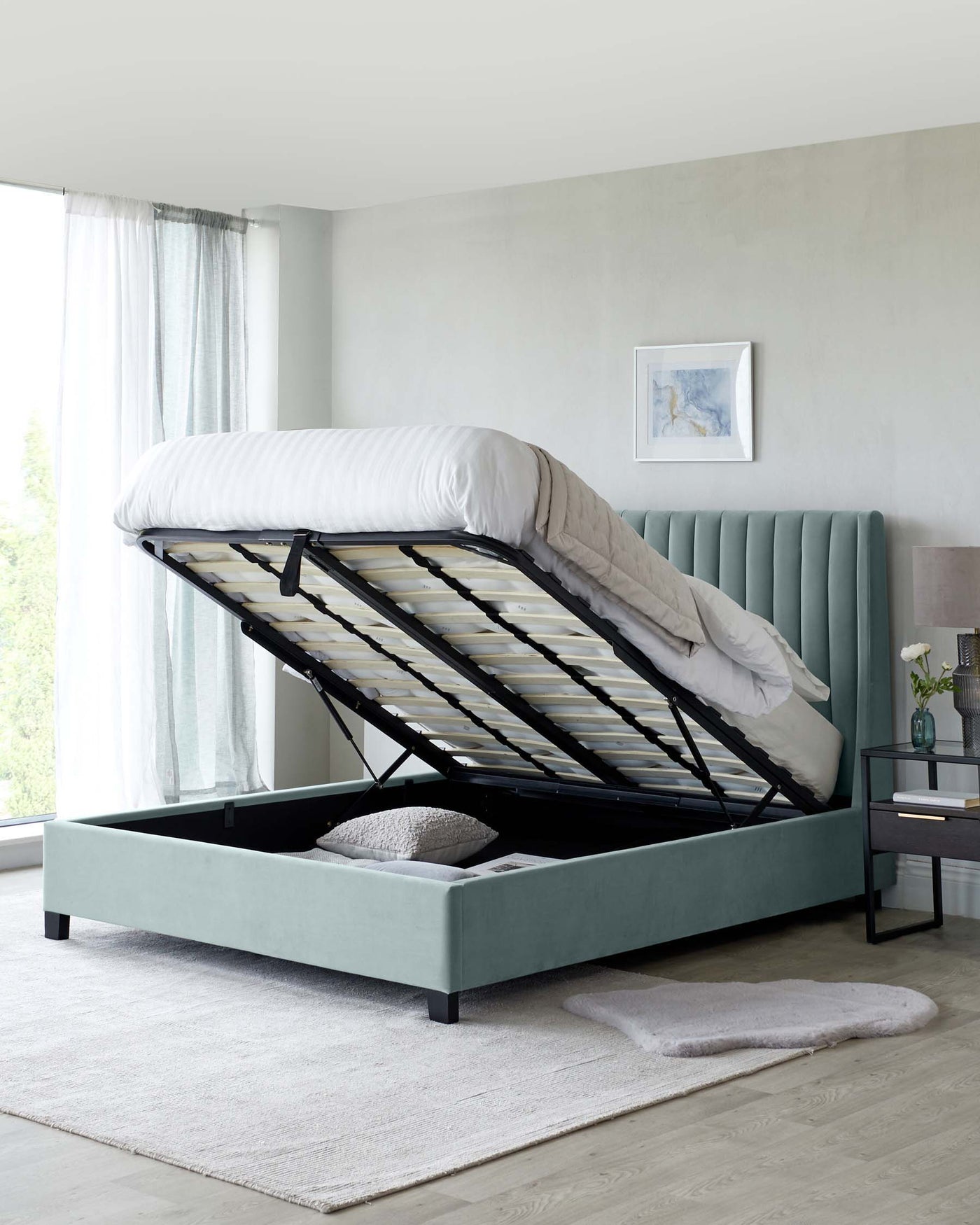 A modern teal upholstered storage bed with the mattress lifted to reveal a spacious storage area beneath. It features a panel-styled headboard and is complemented by a sleek black and glass bedside table.