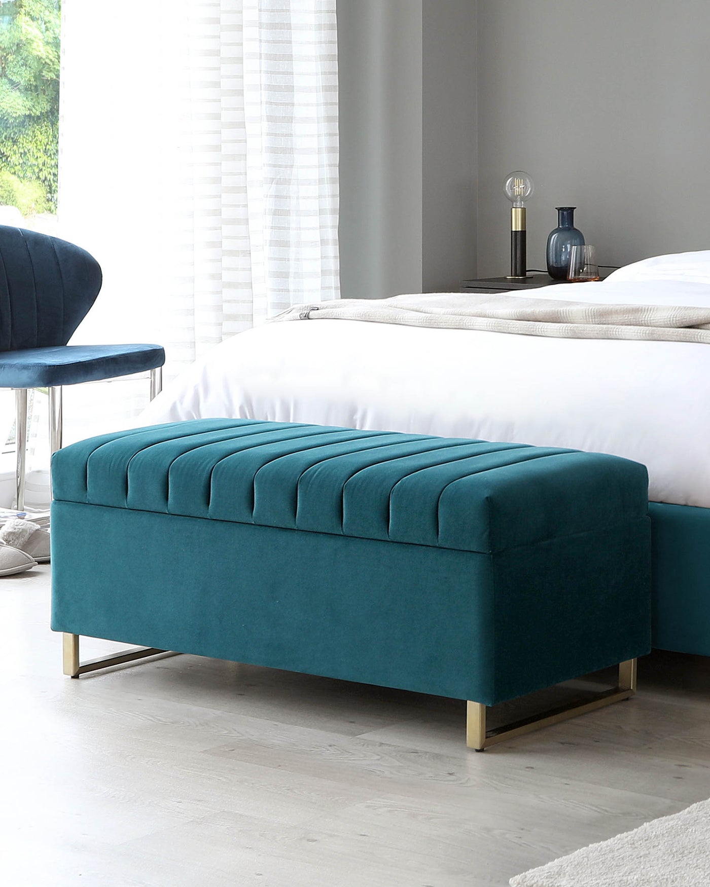 Elegant teal blue upholstered bench with plush, channel-tufted padding and sleek gold-toned metal legs at the foot of a neatly made bed; contemporary design with a luxurious appeal.