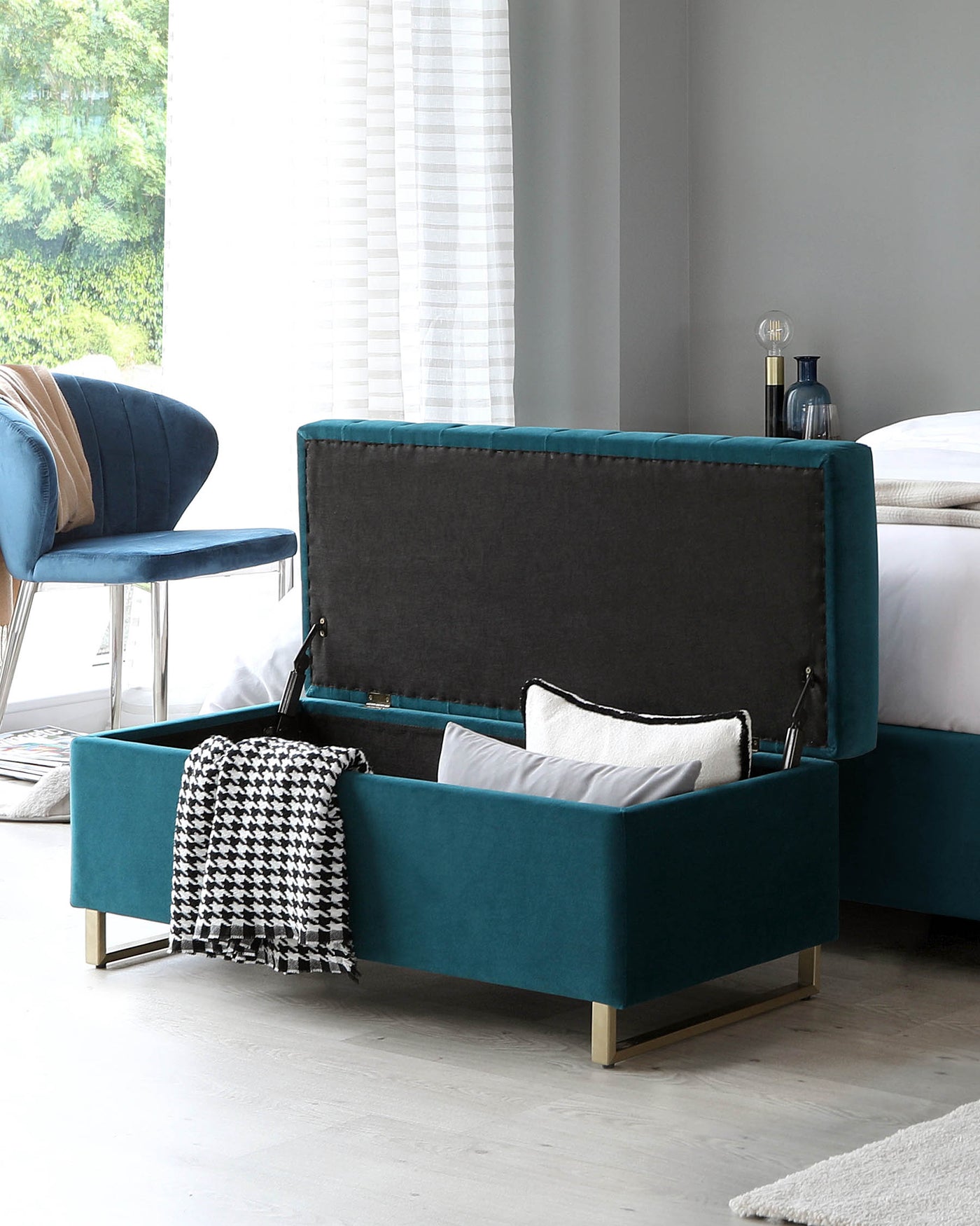 Contemporary style storage bench in teal velvet upholstery with a hinged top, featuring a tufted exterior in a contrasting dark shade, accented by elegant gold-tone metal legs. A black and white patterned throw blanket is casually draped over one side.