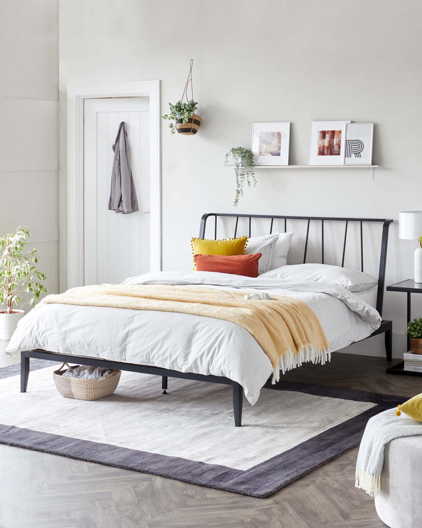 Modern minimalist black metal bed frame with clean lines and a matching black bedside table featuring a round tabletop and minimalistic frame. A white ottoman with tufted details is seen at the foot of the bed. The bed is complemented by soft white and beige bedding with colour-accented throw pillows.