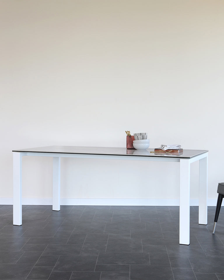Modern minimalist white dining table with a sleek surface and straight legs, paired with a black stool with thin metal legs. Accessories on the table include a decorative vase and a stack of books, emphasizing the table's spacious, uncluttered design.