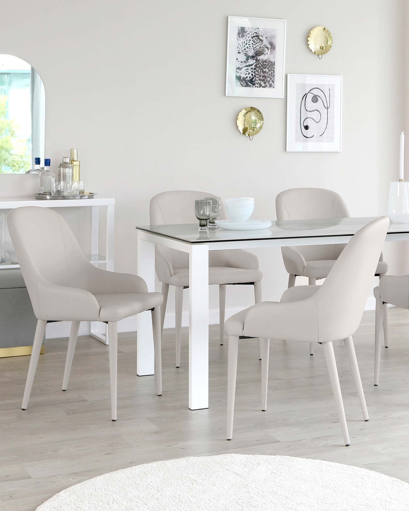 Modern minimalist dining set featuring a white rectangular table with a glass top and sleek metal legs, accompanied by four upholstered chairs in light grey fabric with slender, tapered legs in a matching white finish. A white console table with decorations stands against the wall in the background.