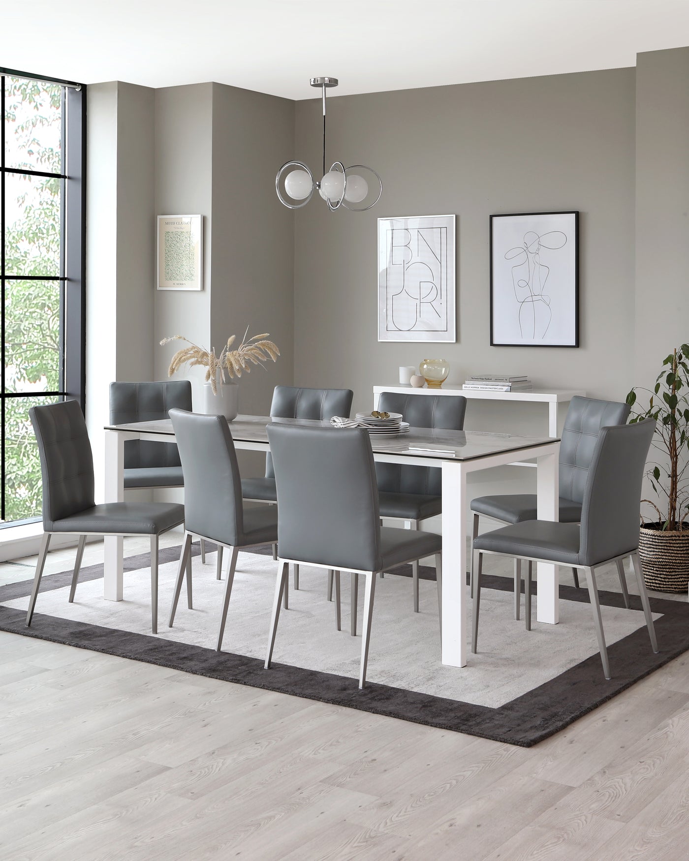 Modern dining room furniture featuring a white rectangular table with stainless steel legs, accompanied by six elegant grey upholstered chairs with sleek metal frames. The set is grounded by a two-tone area rug.