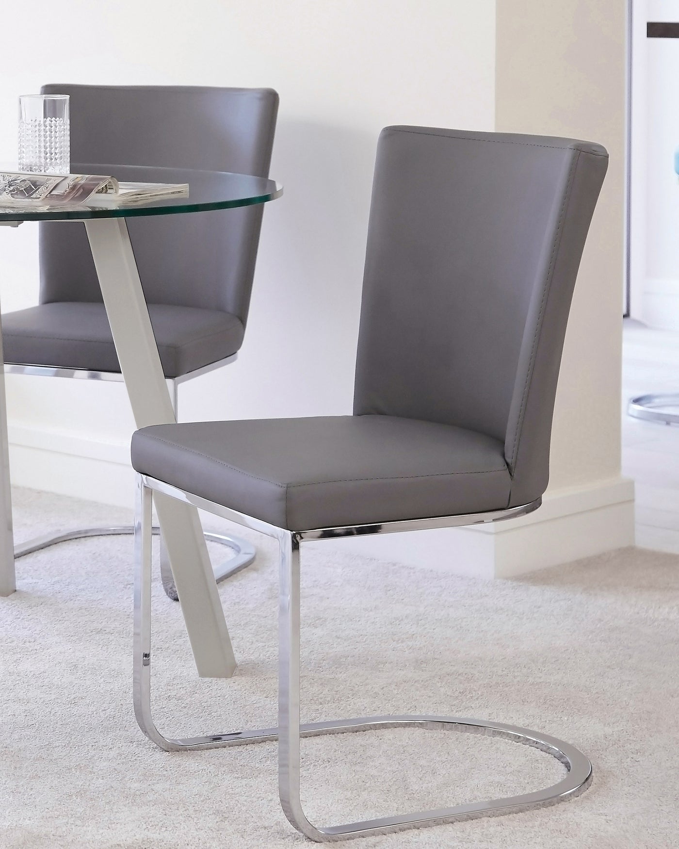 Modern grey upholstered dining chair with a rectangular high backrest and cantilever chrome base, alongside a round glass table with a single chrome leg support.
