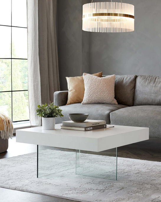 A modern minimalist rectangular white coffee table with a smooth finish and clear glass legs, complemented by a plush grey sofa with soft throw pillows, displayed in a contemporary living room setting. A light grey rug anchors the space.