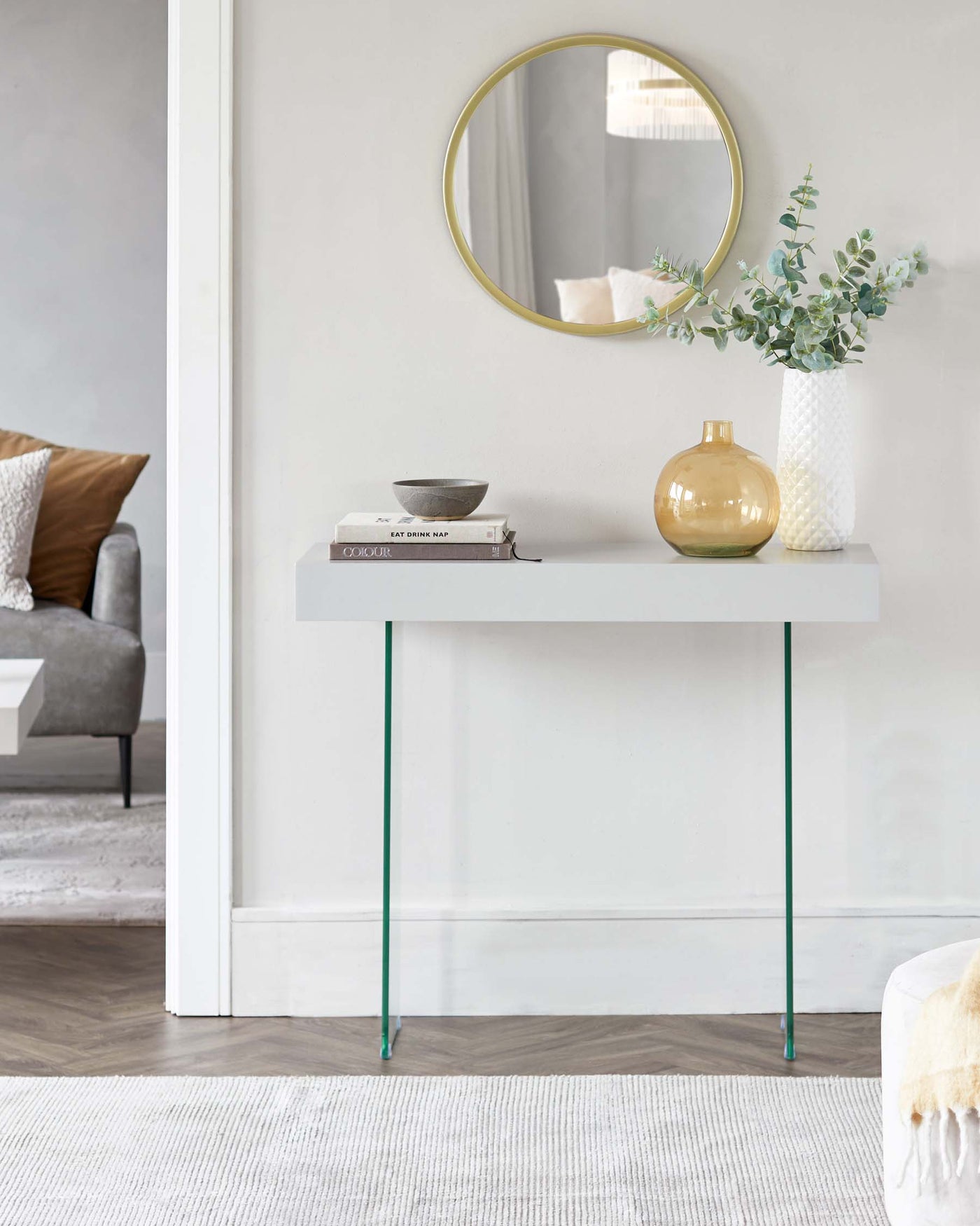 A modern minimalist white console table with slender green metal legs, adorned with decorative items including books, a bowl, a vase, and a round gold-framed mirror hanging above it.