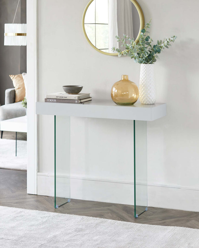 Modern minimalist white console table with slender green-tinted glass legs, styled with decorative books, a textured ceramic vase with foliage, and a spherical amber glass vase. The table is set against a light neutral wall beside a round mirror.