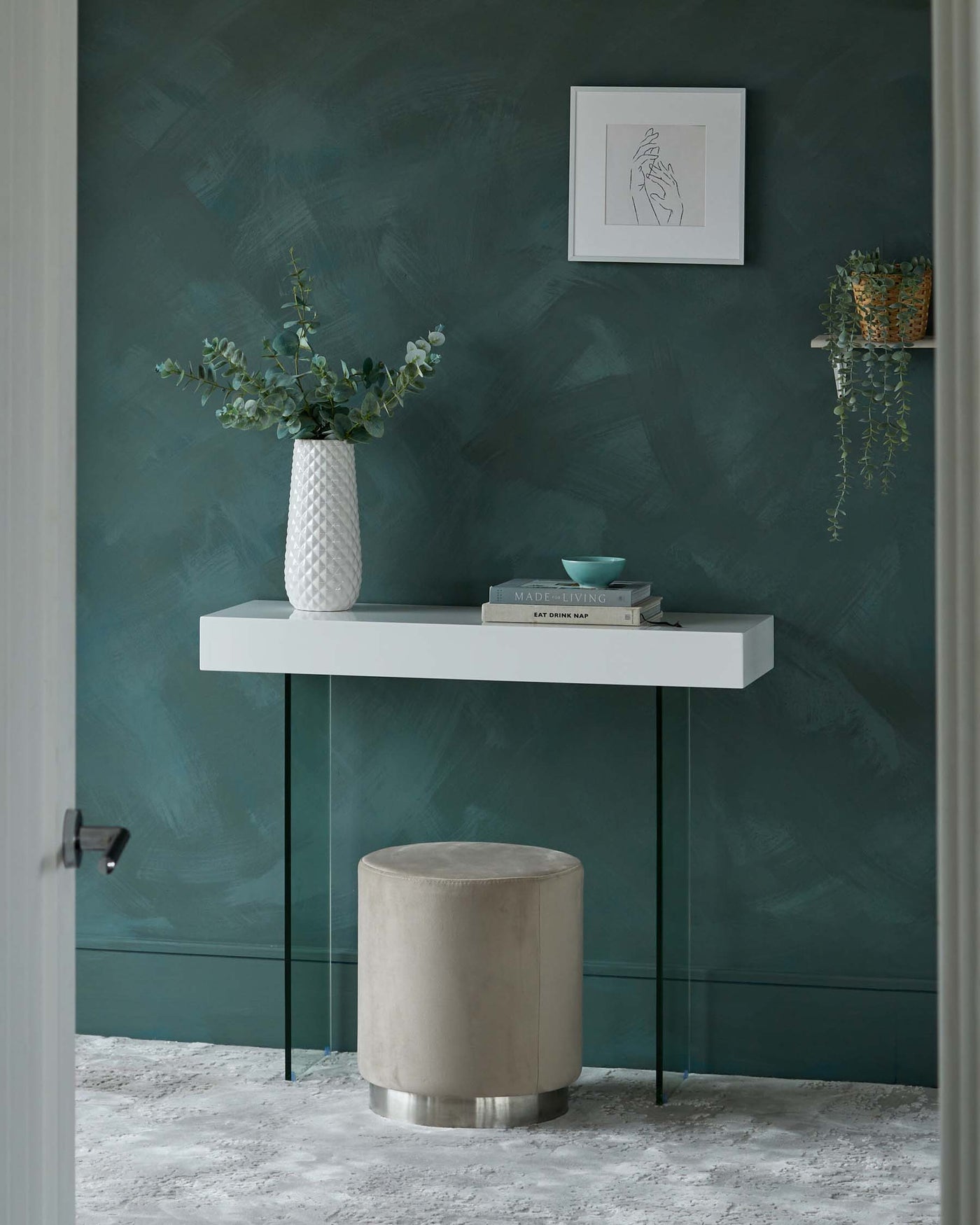 Modern minimalist floating wall console table with a sleek white top and two metal support legs in black finish, paired with a cylindrical light-beige velvet upholstered ottoman with a silver metallic base, staged on a textured light grey rug.
