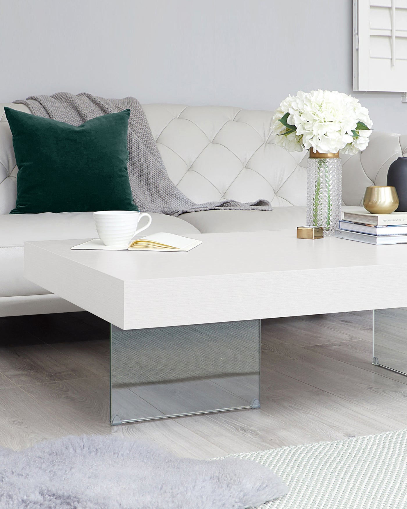 Modern living room featuring a white tufted sofa with dark green and grey throw pillows, and a low-profile white coffee table with a sleek design and glass base. A decorative vase with white flowers, cup and saucer, and stack of books adorn the tabletop.