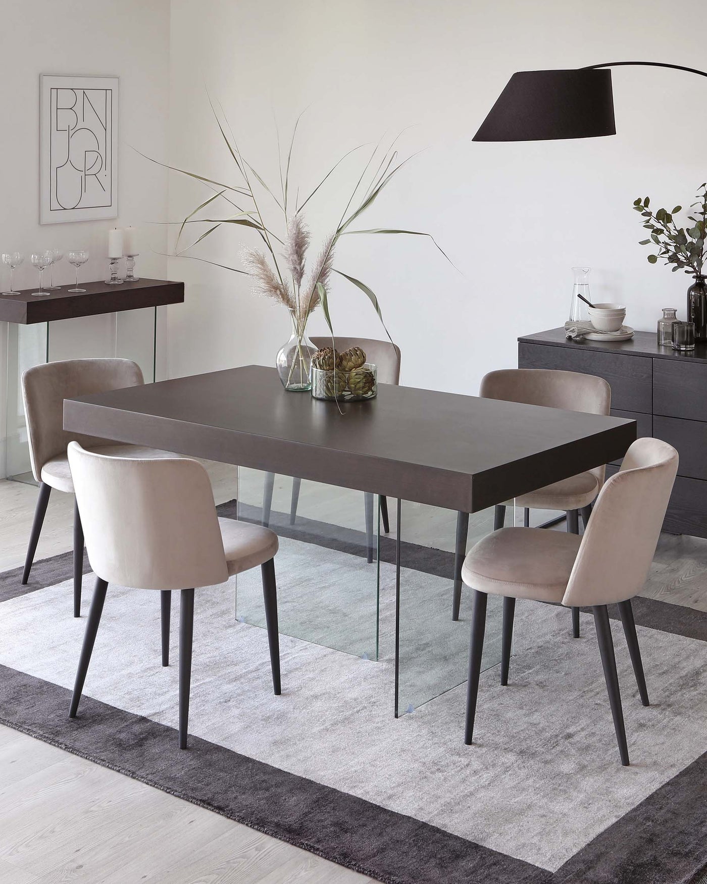A modern dining room set composed of a rectangular dark brown table with glass legs and four upholstered chairs with a velvety beige finish and black legs. There is a sideboard in the background matching the table, and a minimalist black floor lamp with an oversized lampshade arches over the scene.
